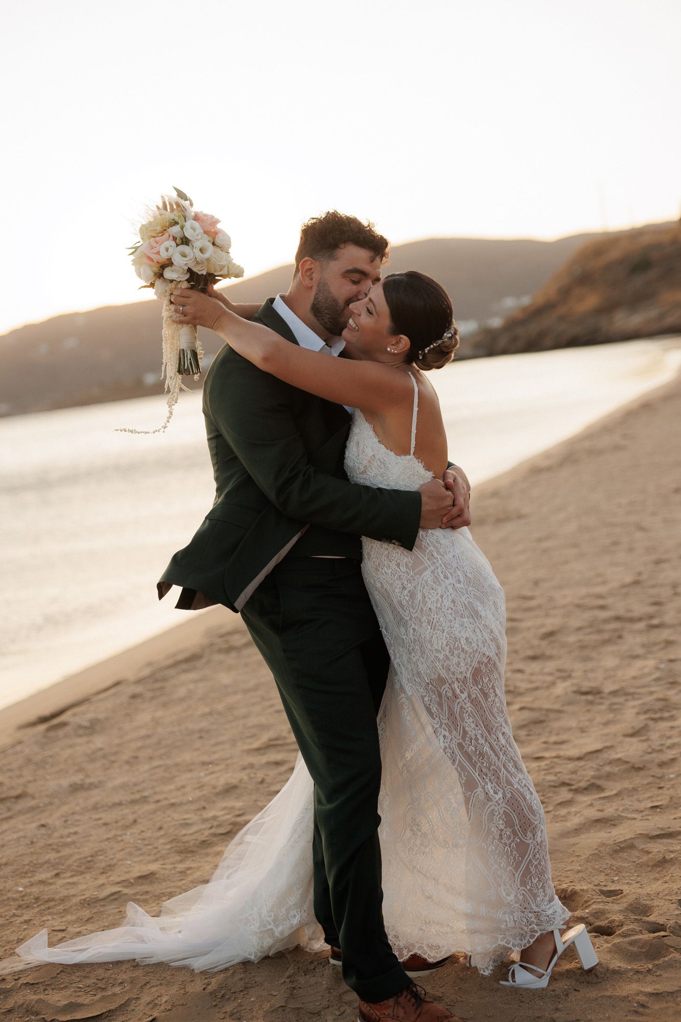 Bride and groom share romantic embrace on sandy beach at sunset holding white bouquet.