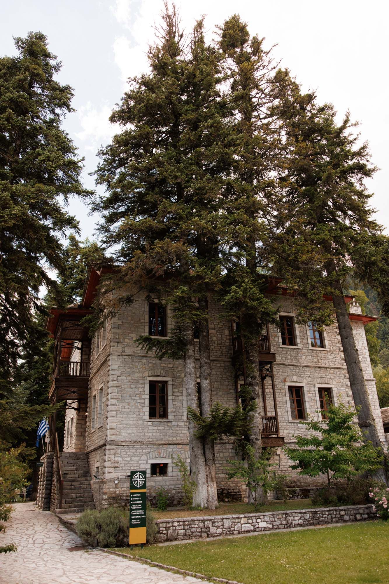 Historic stone building surrounded by tall evergreen trees with Greek flag at entrance.