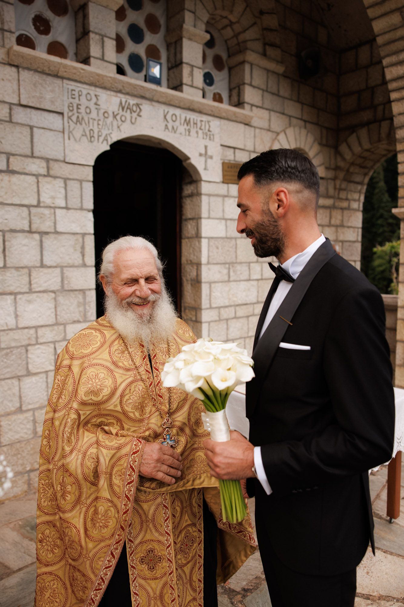 Elderly Orthodox priest in golden vestments greeting a groom holding white calla lily bouquet.