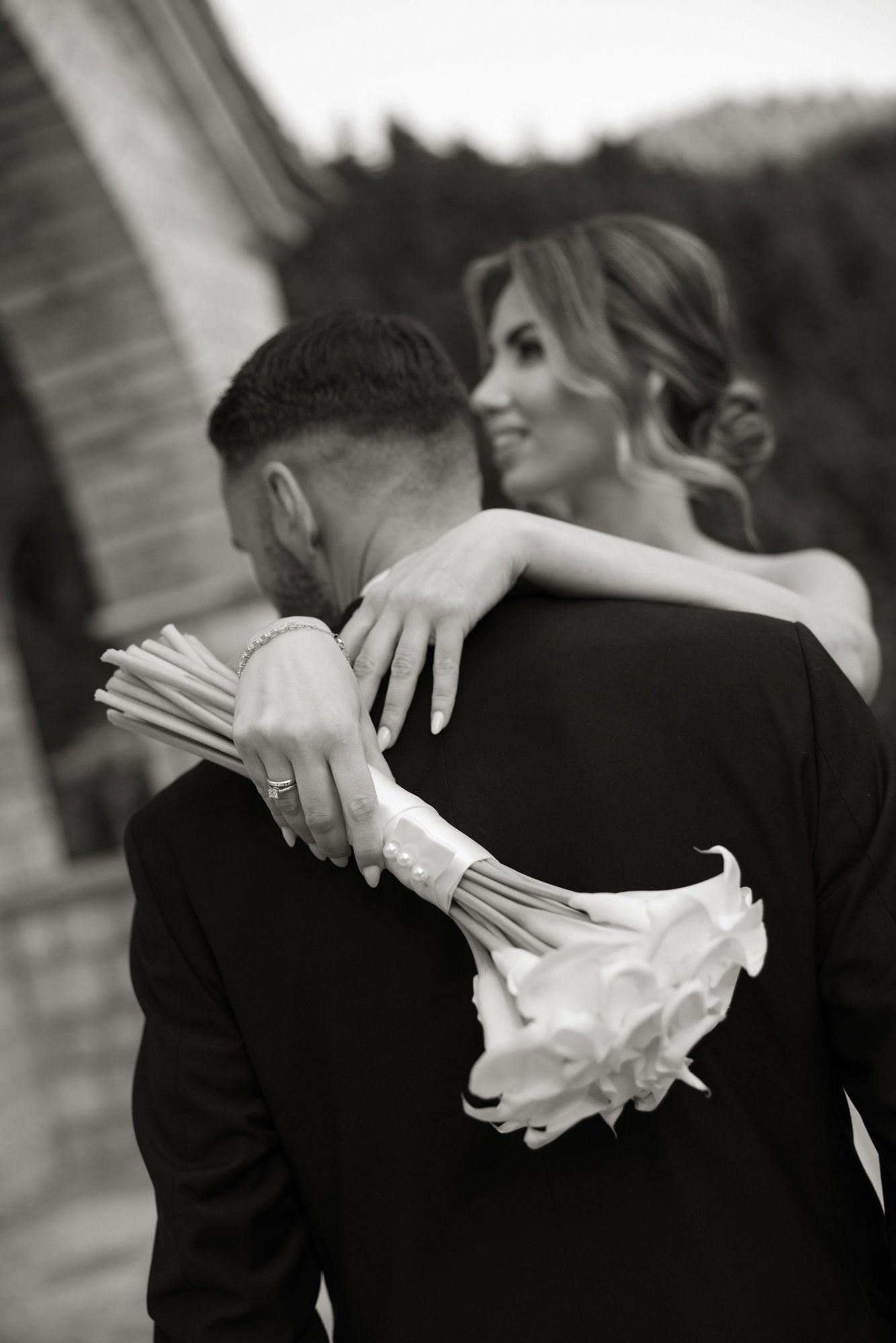 Bride embraces groom from behind holding white calla lily bouquet in black and white wedding photo.