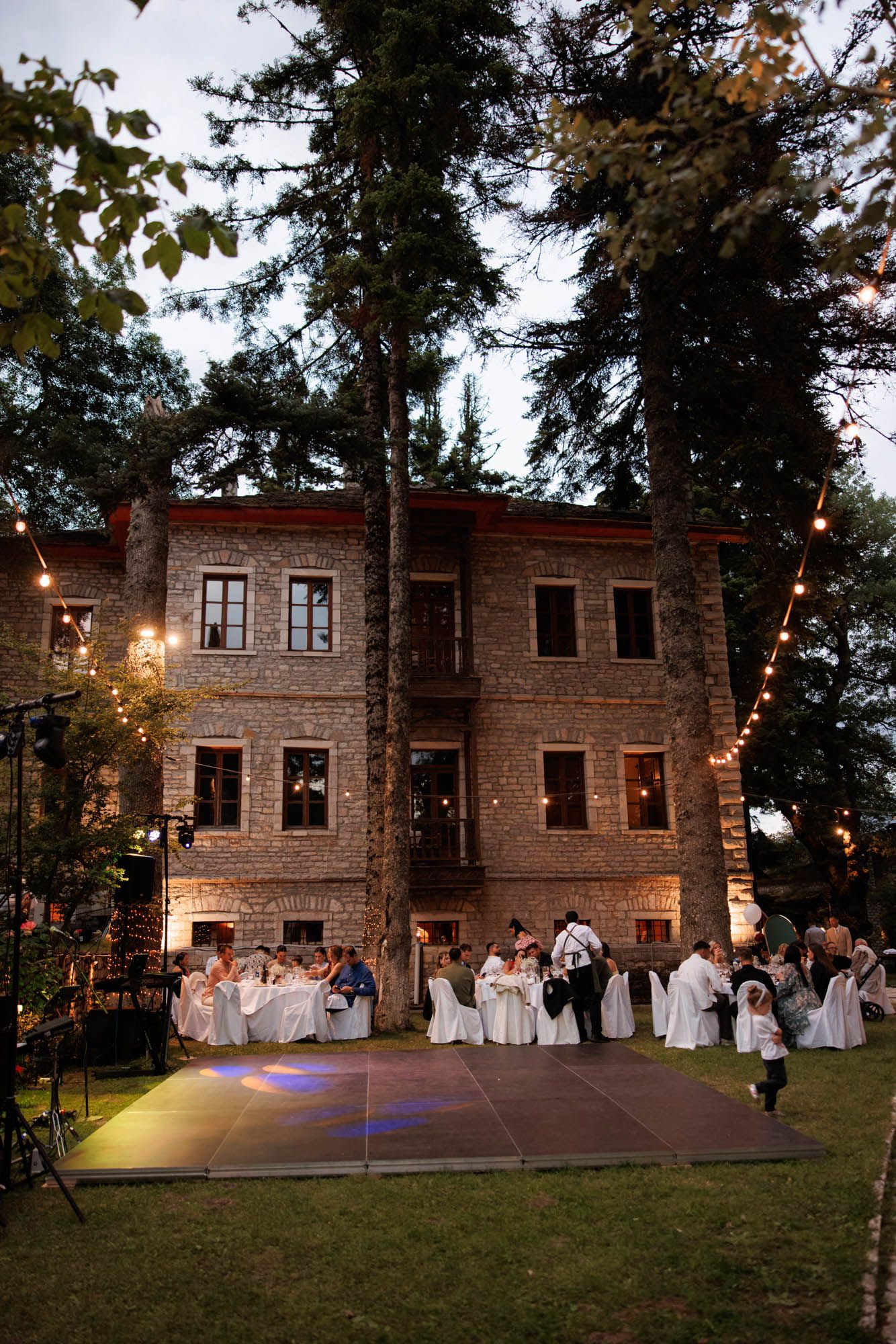 Elegant outdoor wedding reception with dance floor and string lights beside illuminated stone manor at twilight.