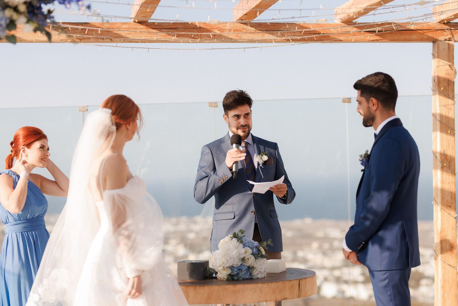 Officiant reads vows at outdoor wedding ceremony under wooden arch with sea view backdrop.