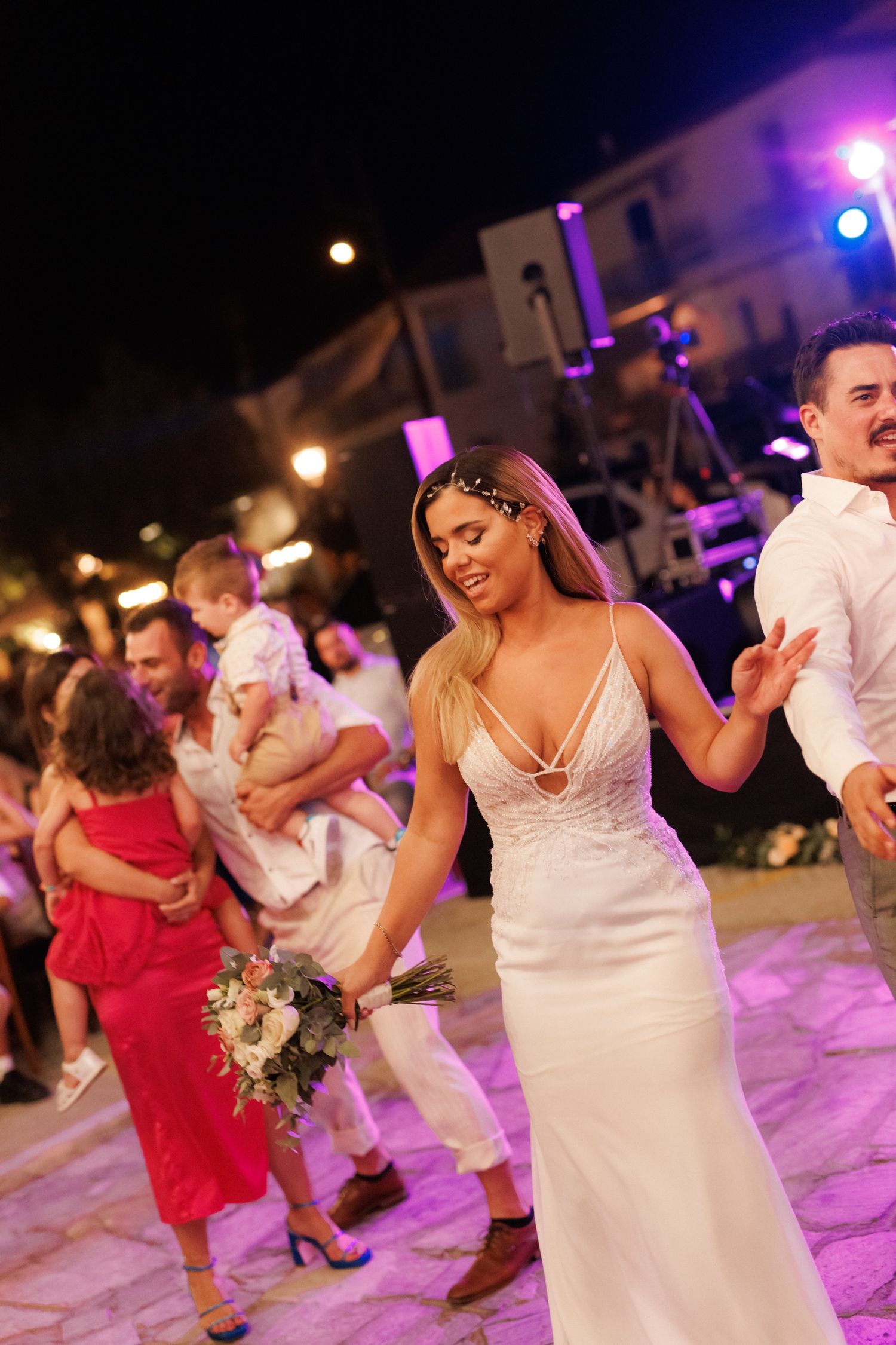 Bride in white gown dancing joyfully with guests at an outdoor wedding reception at night.