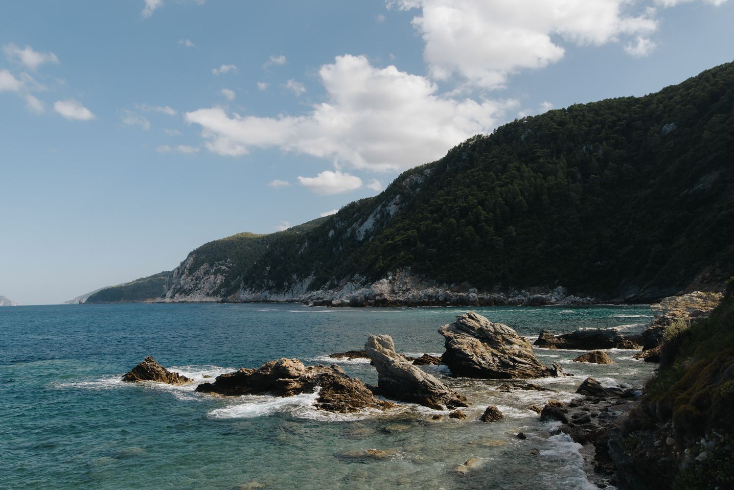 Scenic coastal landscape with rocky shoreline, turquoise sea, and lush green hillside under blue sky.