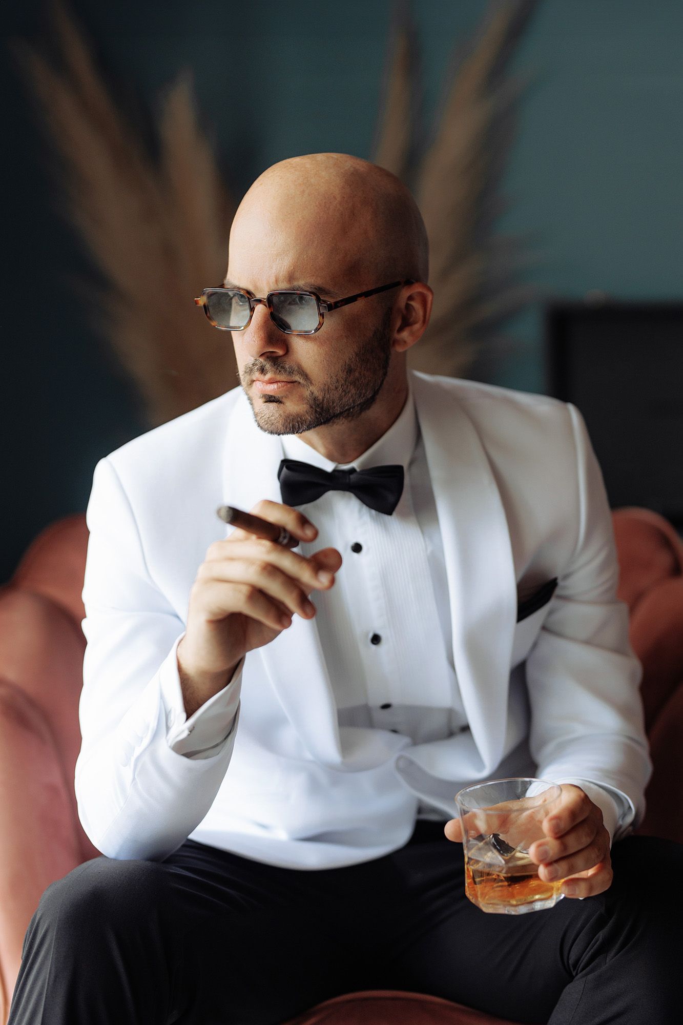 A person in a white tuxedo and bow tie sits on a pink chair holding a cocktail and cigar.