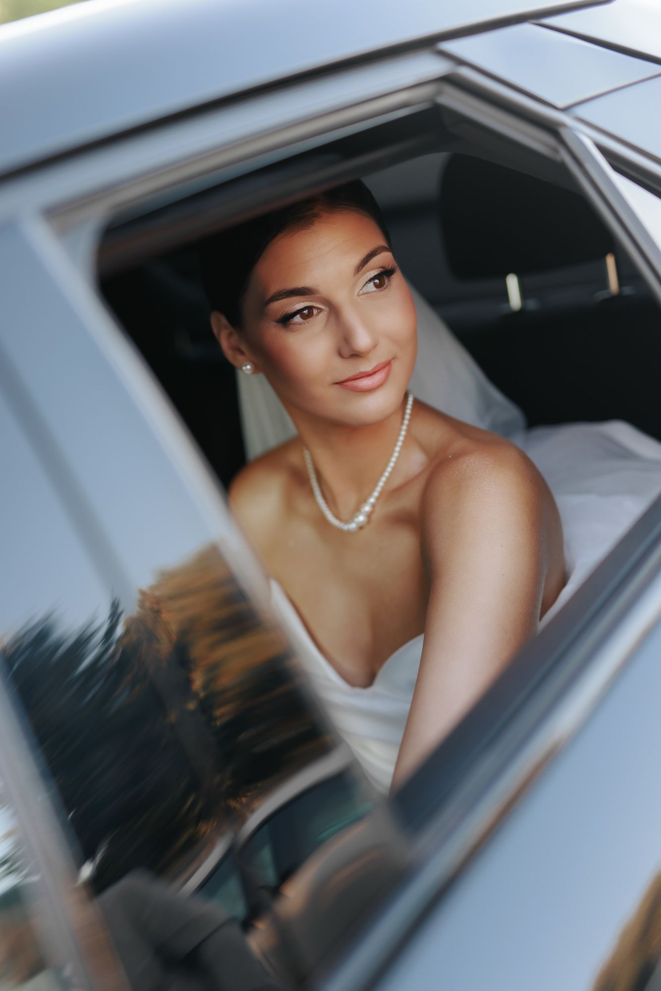 Elegant bride in white dress and pearl necklace gazing from a car window on her wedding day.