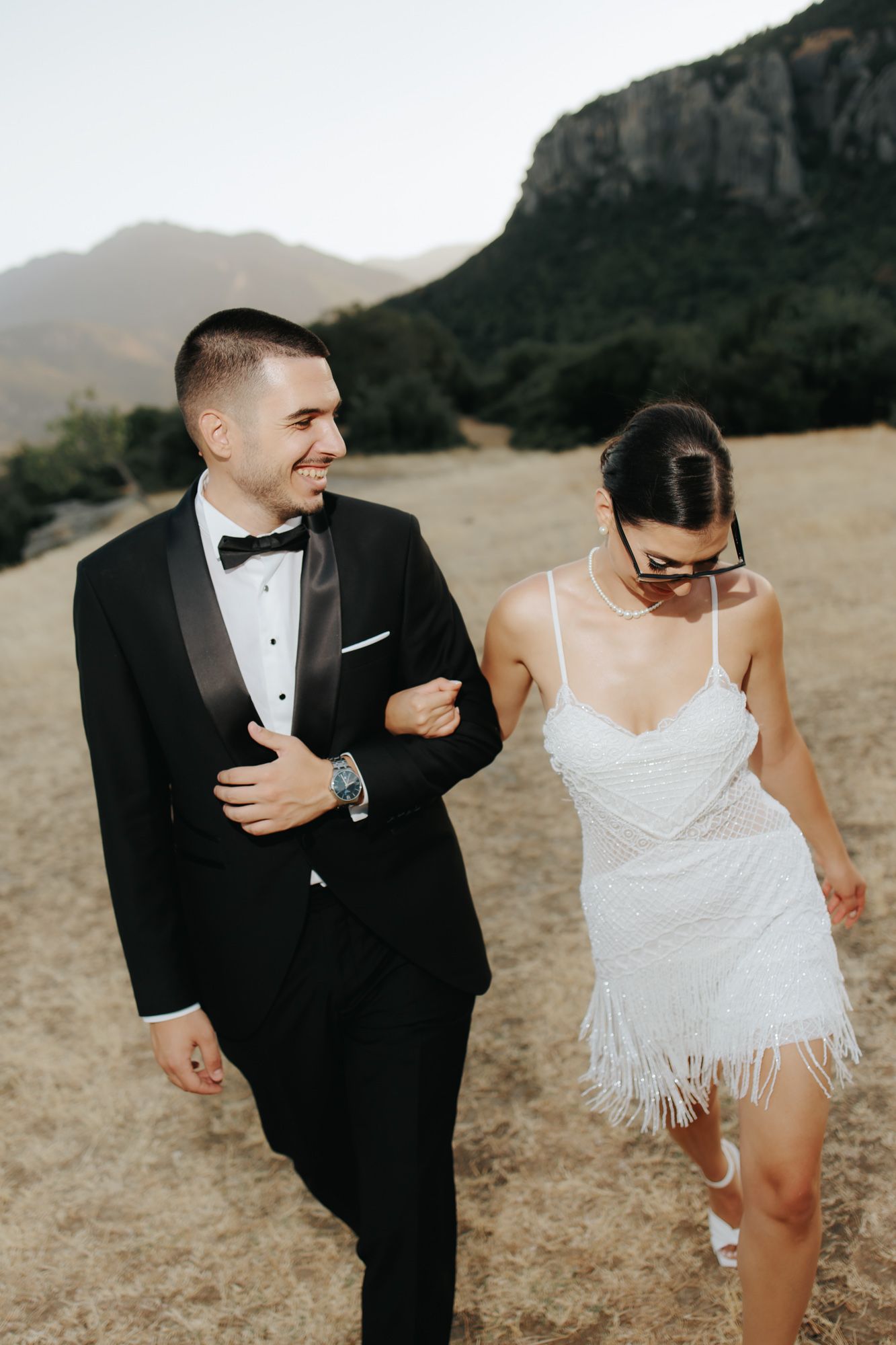 Bride in white fringed mini dress and groom in black tuxedo walking together with mountain backdrop.
