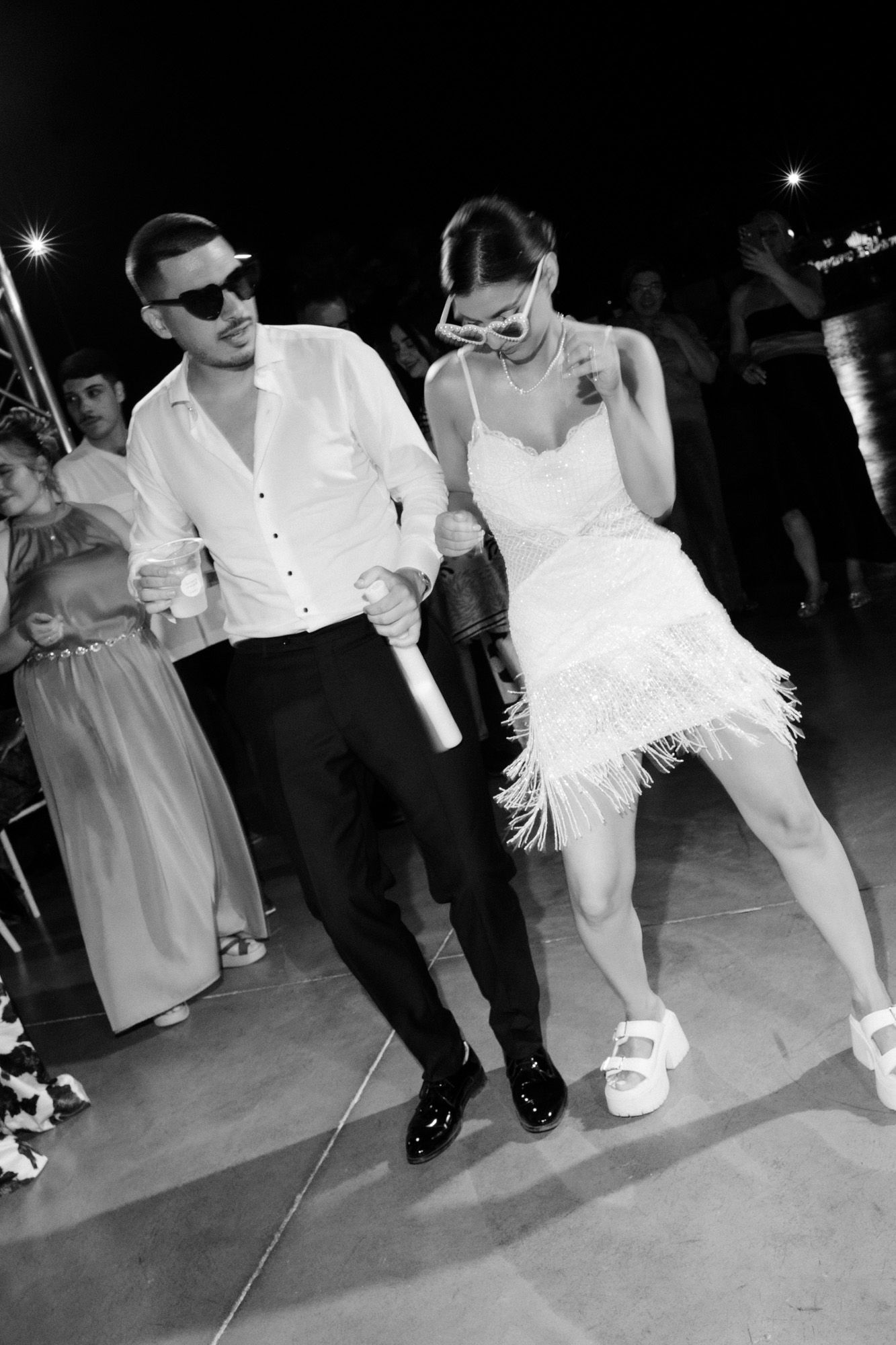 Bride and groom dancing joyfully at wedding reception in black and white photo, wearing sunglasses and white outfits.