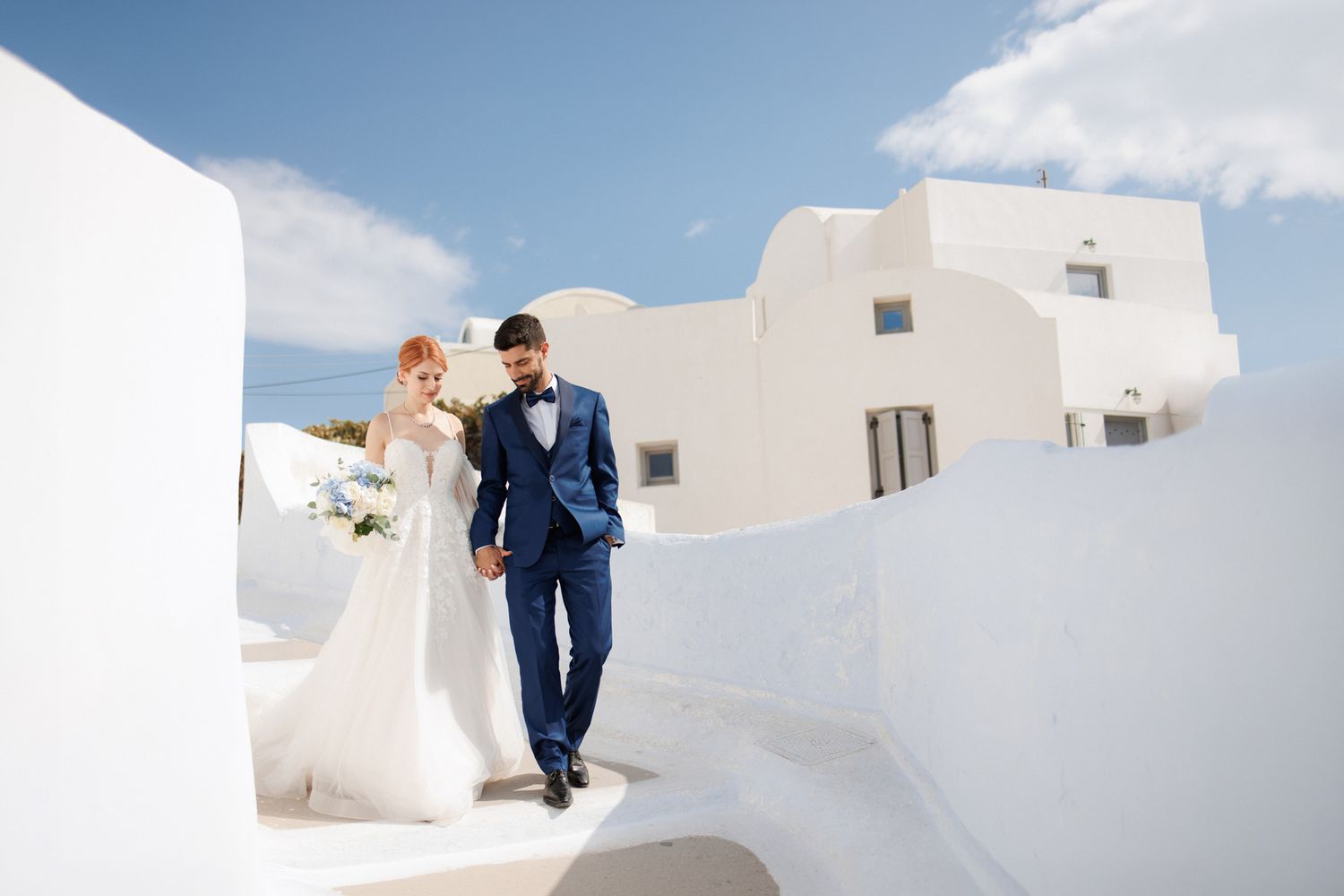 Bride and groom walking hand in hand through white-washed Santorini architecture under a bright blue sky.