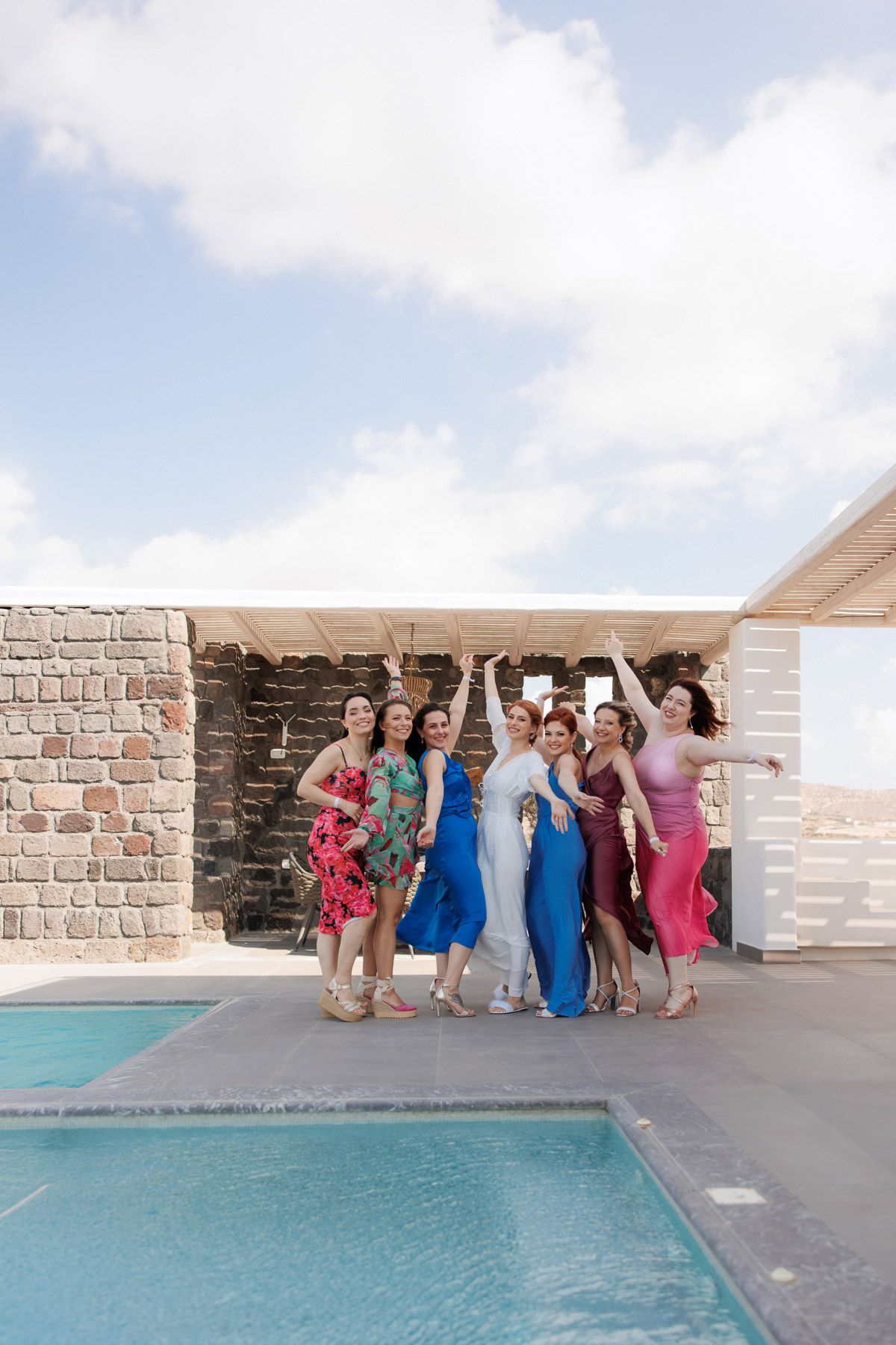Bride and bridesmaids in colorful dresses celebrating joyfully beside a pool at a Greek island villa.