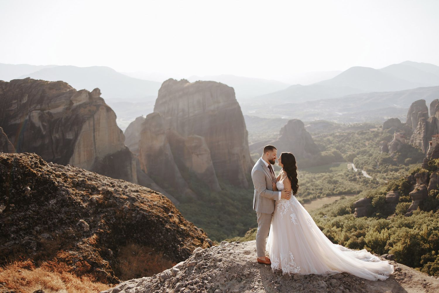 A couple in wedding attire embraces on a rocky cliff overlooking the dramatic mountain landscape of Meteora, Greece.