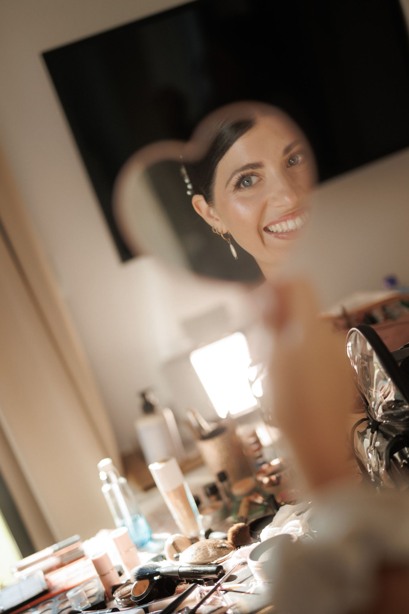Happy bride smiling at mirror reflection during makeup application at a well-lit vanity table.