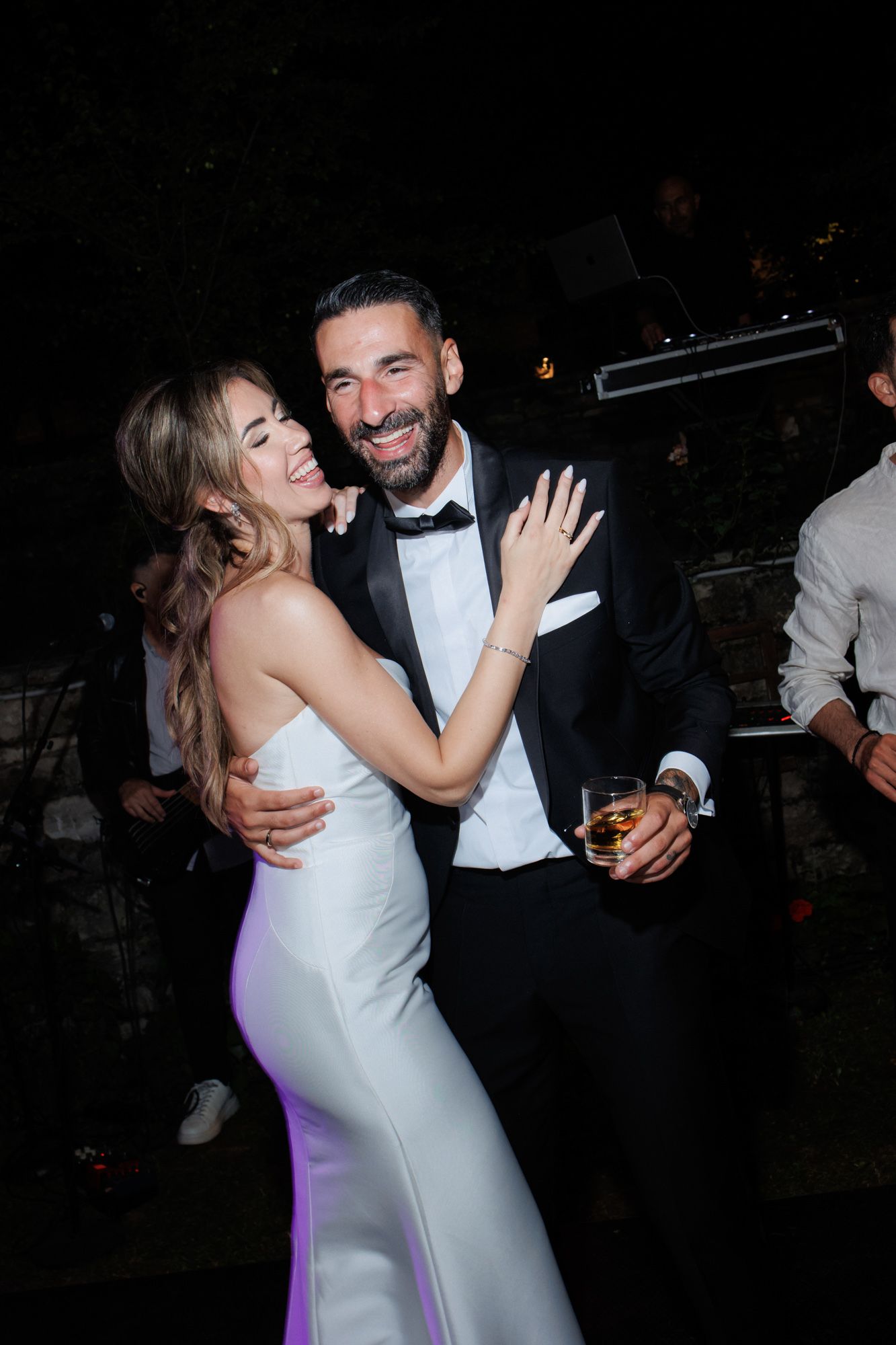 Bride in white strapless gown embracing smiling groom in tuxedo at outdoor wedding reception.