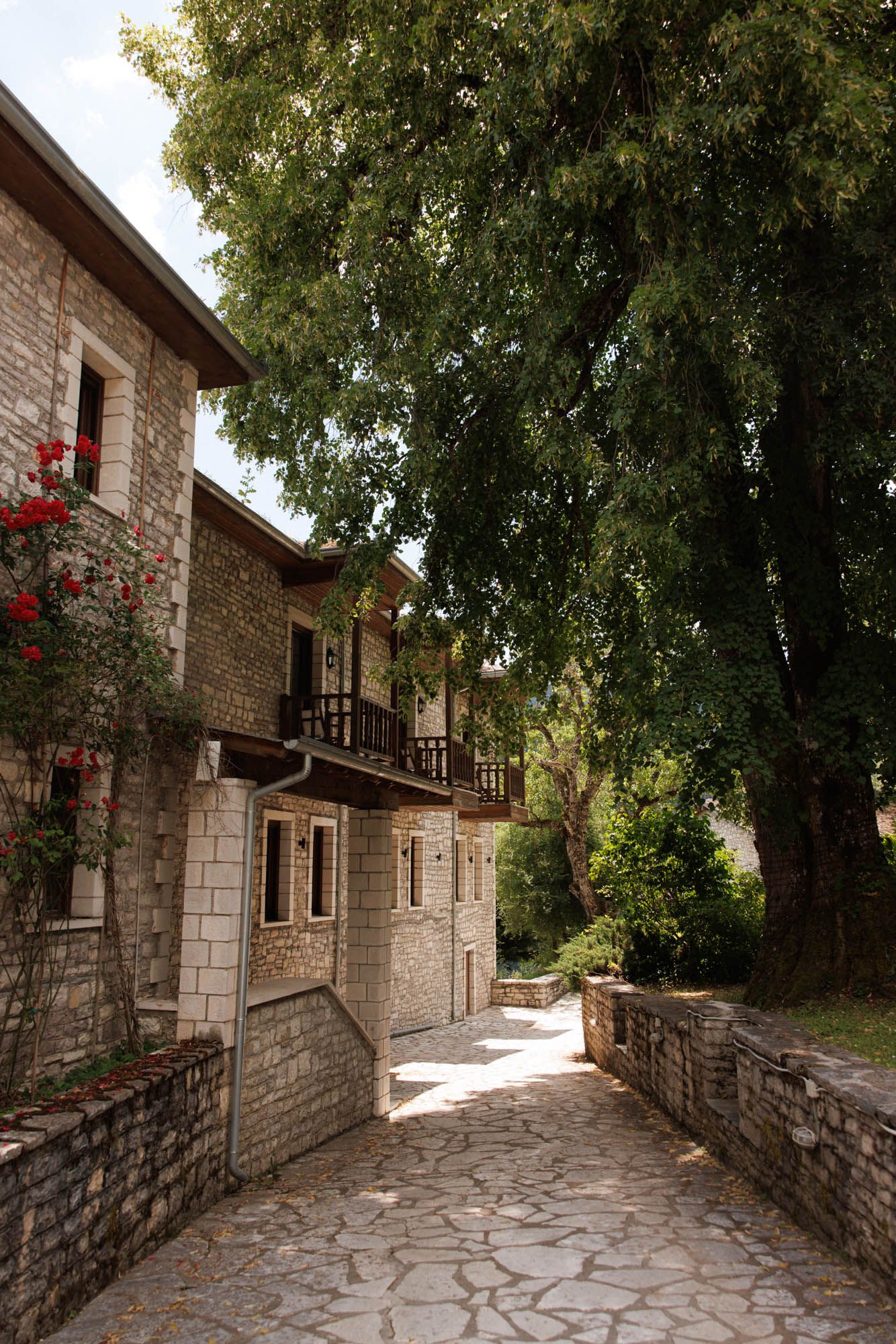 Charming stone building with wooden balcony along cobblestone pathway lined with lush green trees.