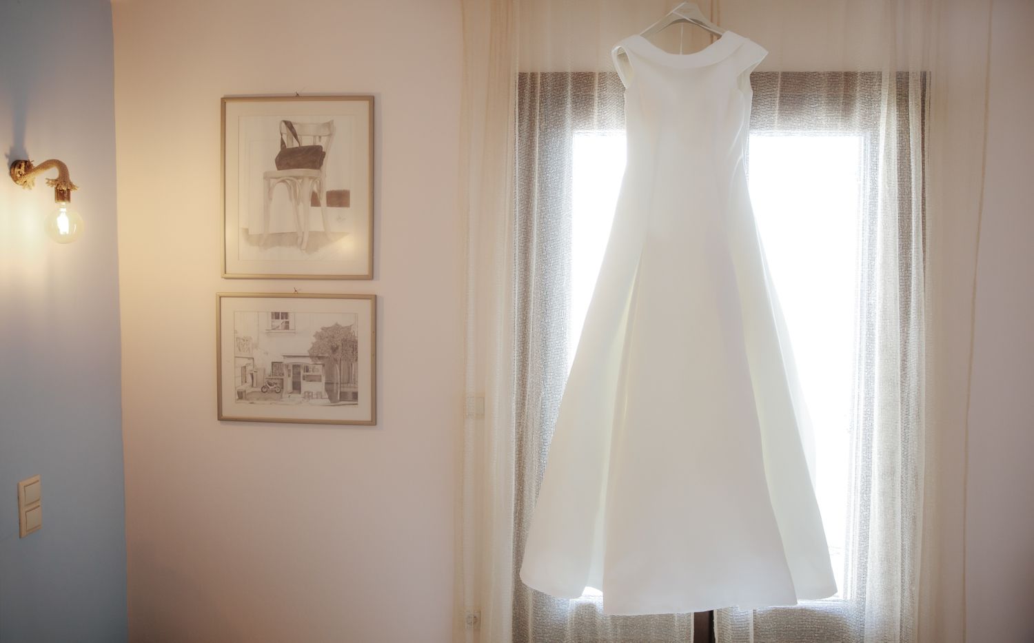 A white wedding dress hanging in a sunlit window with framed artwork on the wall beside it.
