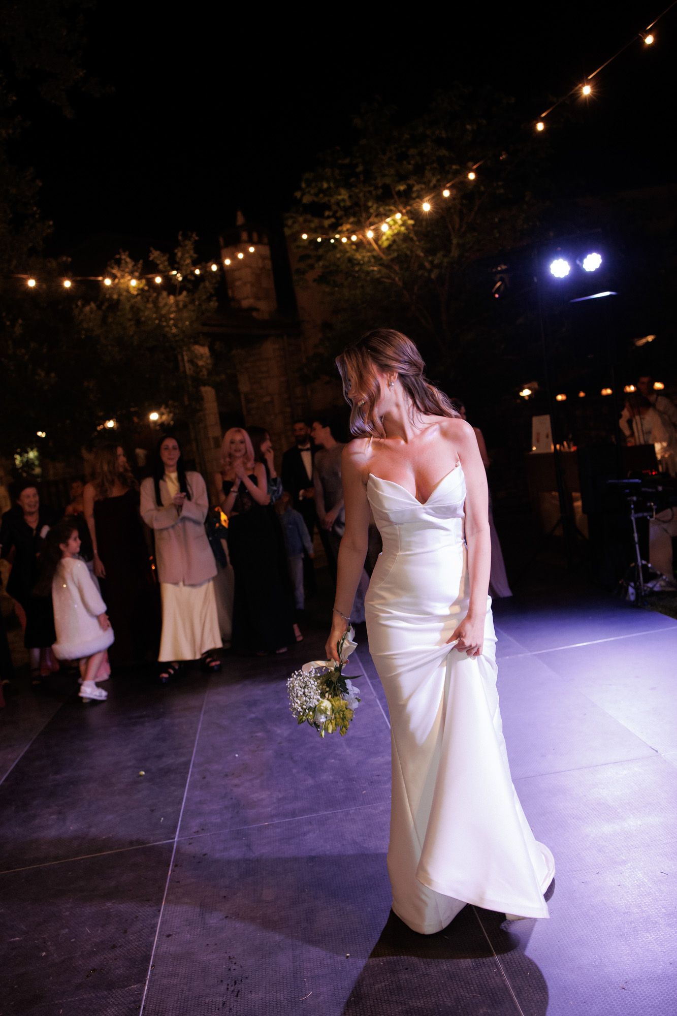 Bride in white spaghetti strap gown holding bouquet on dance floor at outdoor wedding reception.