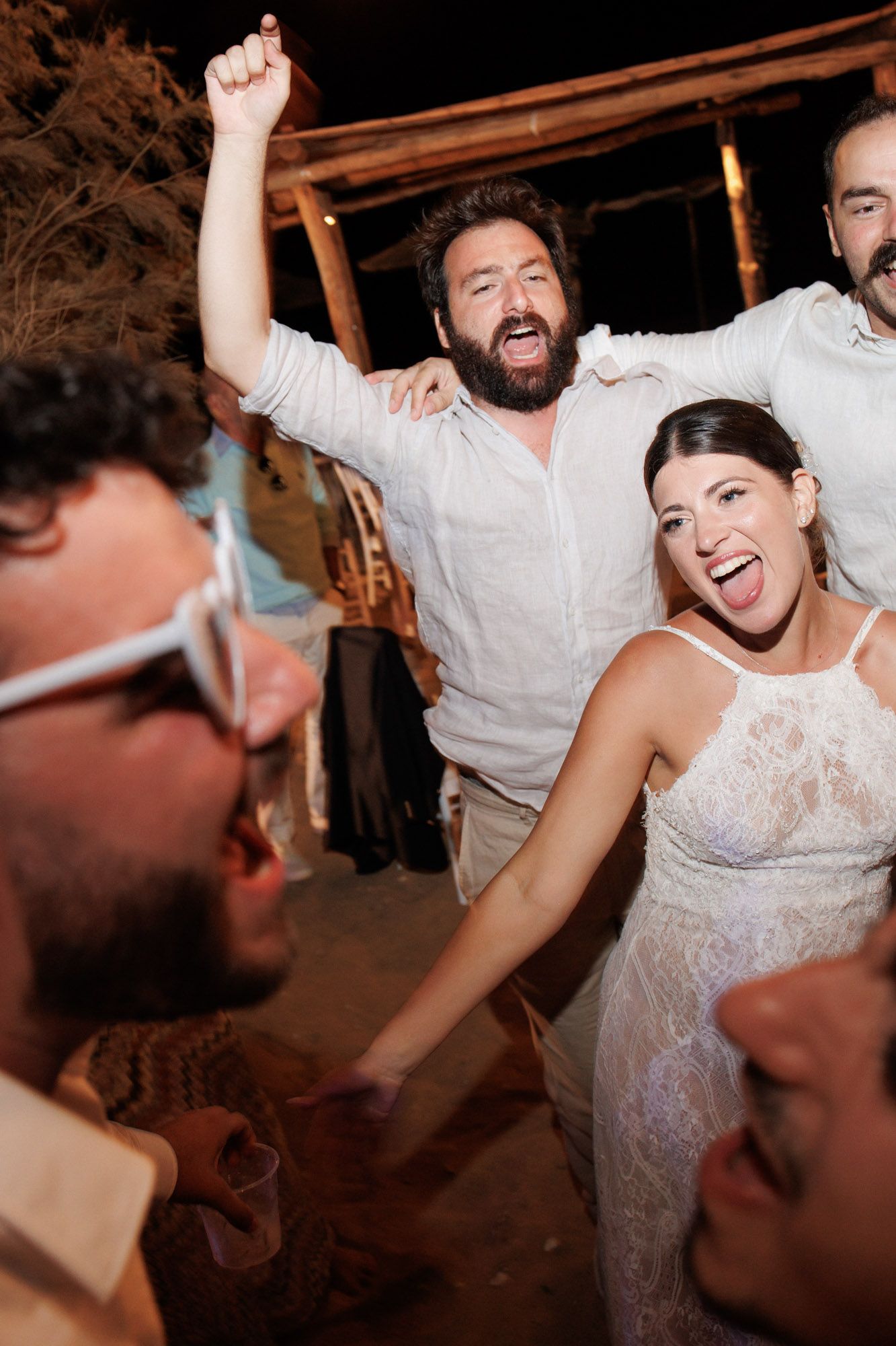 Bride and bearded guest celebrate with raised fists at lively outdoor wedding reception.