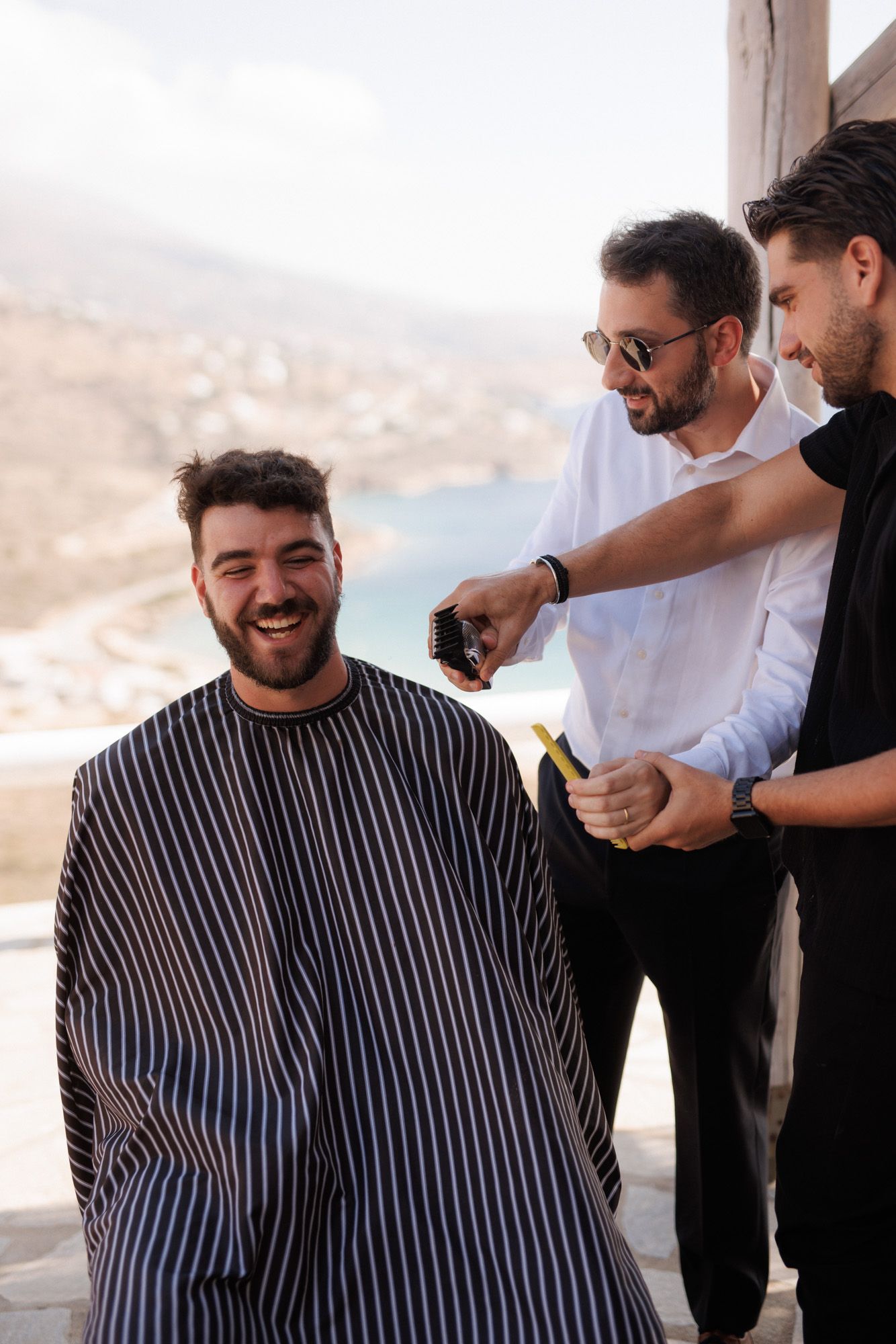 Groom laughing while getting haircut outdoors with scenic Greek island coastline in background.