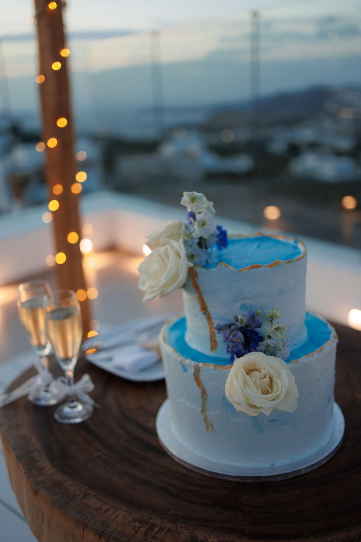 Two-tier white and blue wedding cake with roses and gold details beside champagne flutes.