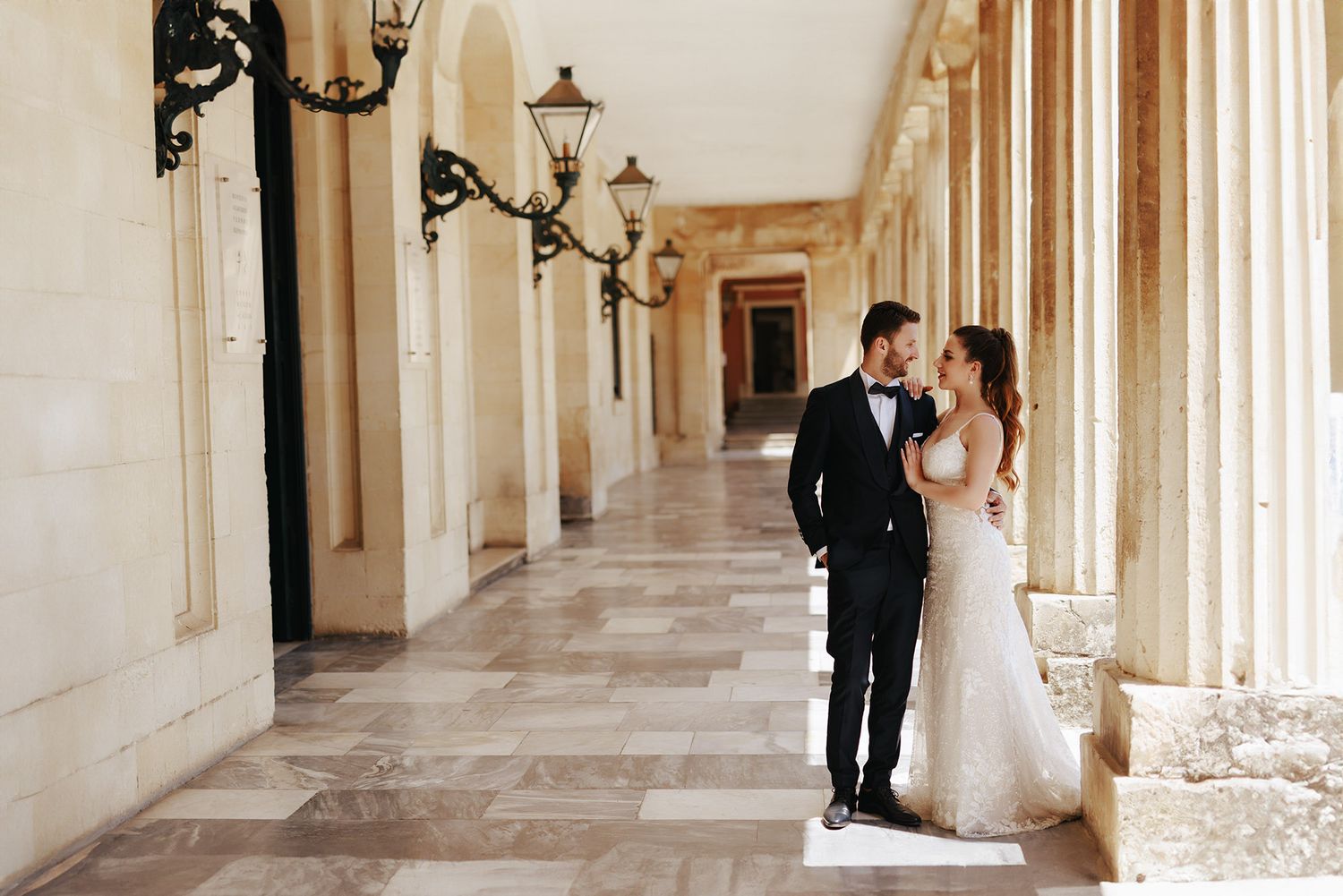 A couple in formal wedding attire standing in an elegant colonnade with ornate lighting fixtures and marble floors.