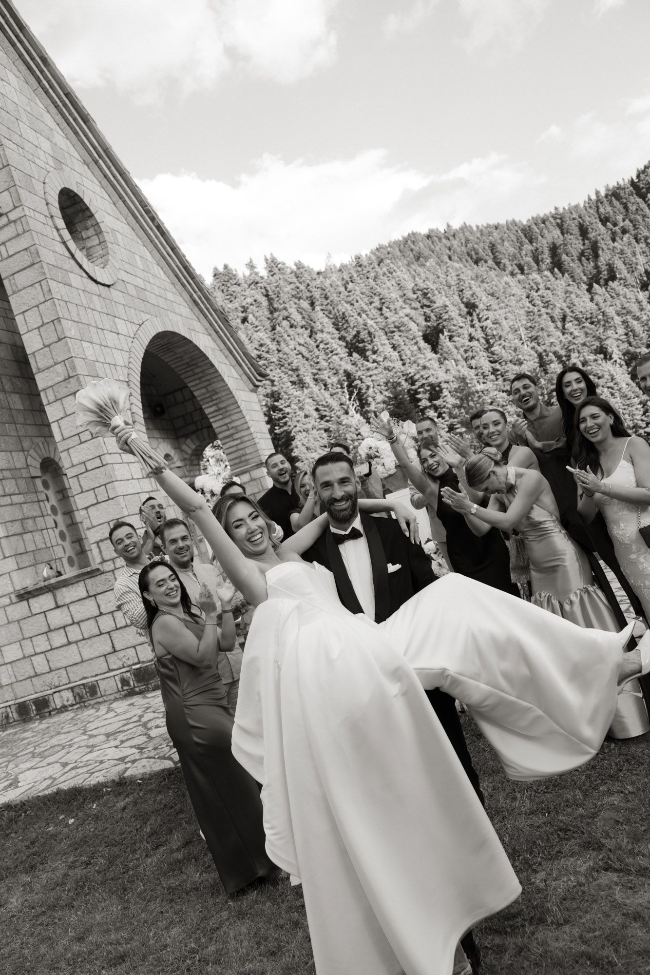 Groom lifts bride surrounded by cheering wedding party outside stone chapel with forest background, black and white.