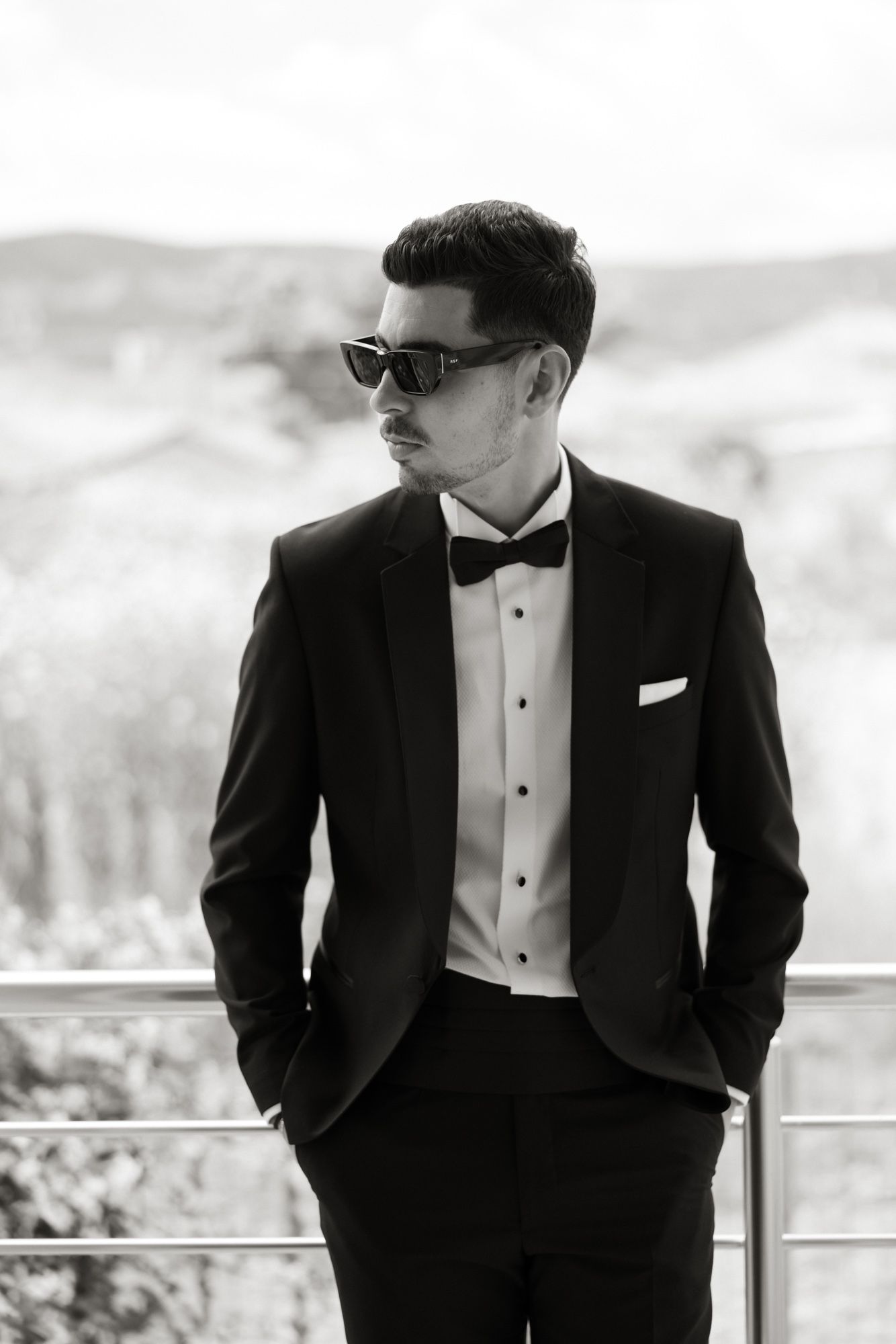 A stylish man in a black tuxedo with bow tie and sunglasses poses confidently outdoors in a black and white photo.