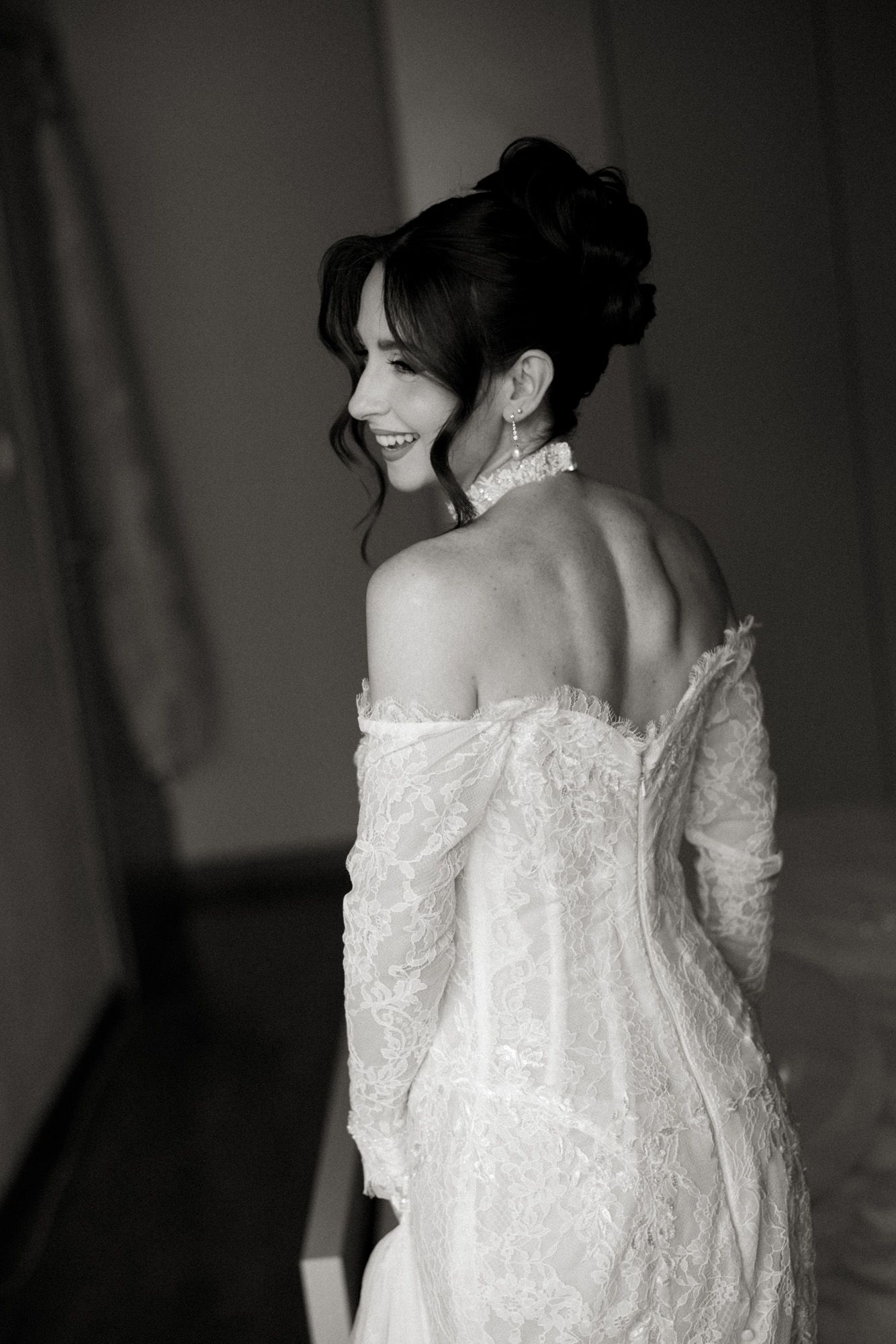 Black and white photo of a smiling bride in an off-shoulder lace wedding gown with elegant updo and drop earrings.