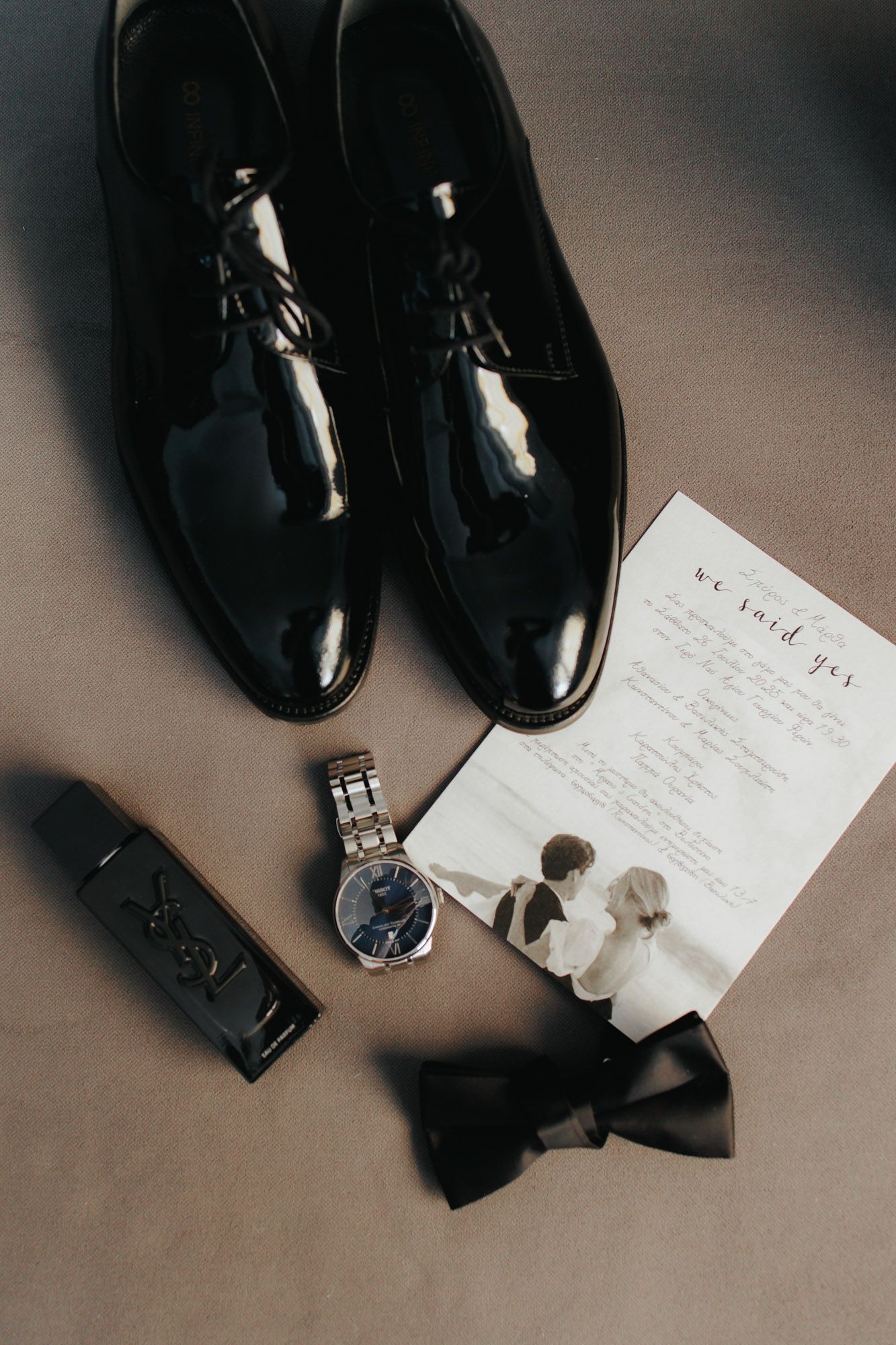 Groom wedding accessories flat lay with black patent shoes, watch, bow tie, YSL cologne, and wedding invitation card.