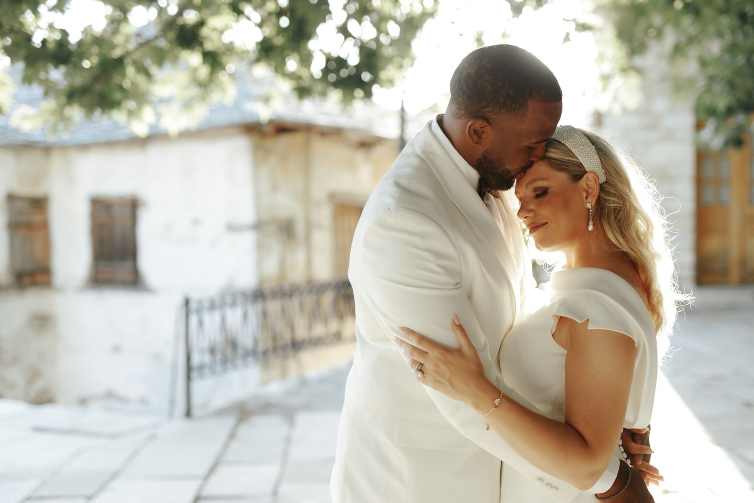 Interracial couple in white wedding attire sharing a romantic embrace outdoors near a sunlit stone building.
