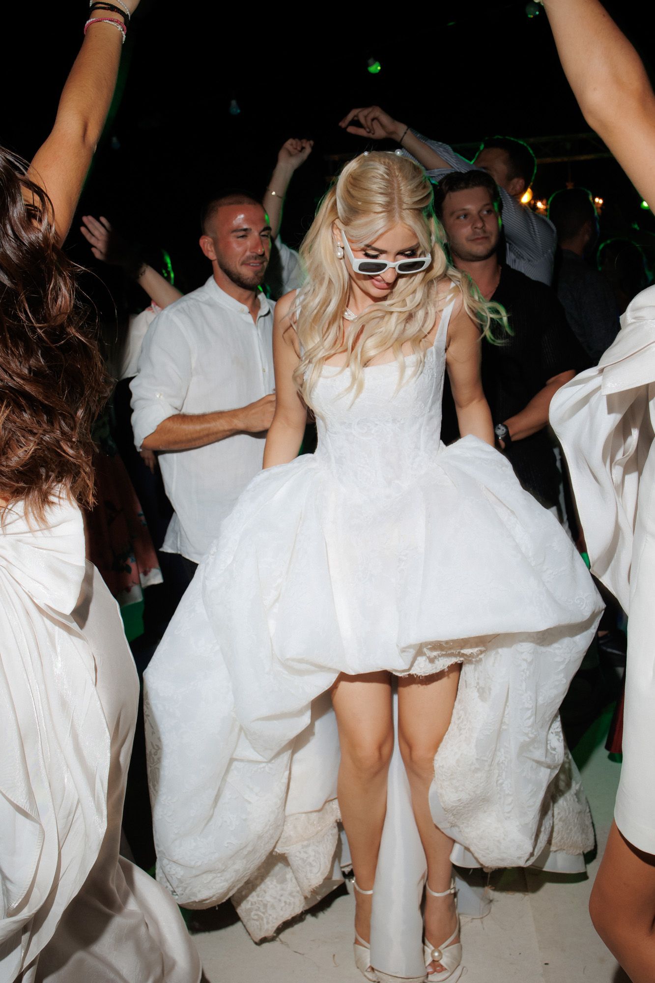 Bride in short white dress and sunglasses dancing joyfully at wedding reception.