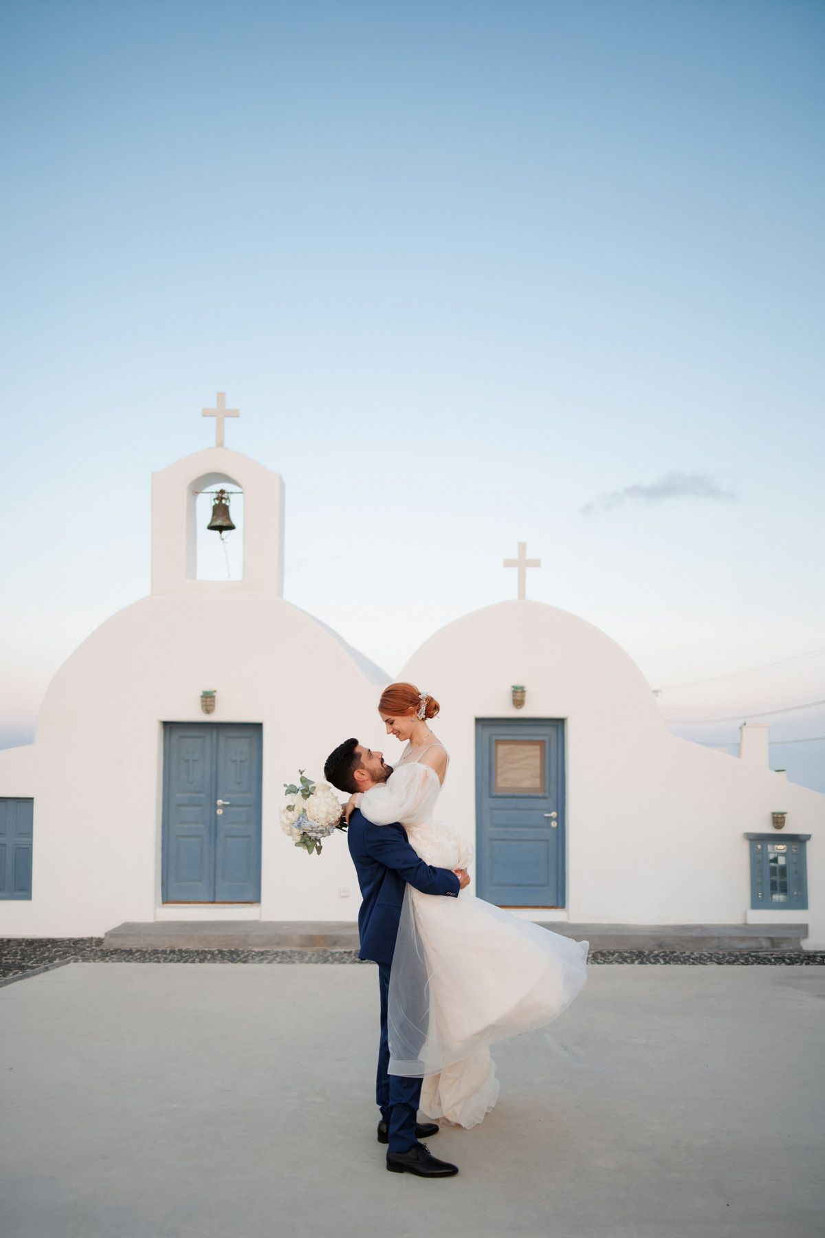 Groom lifts bride in front of white Greek Orthodox church at sunset in Santorini.