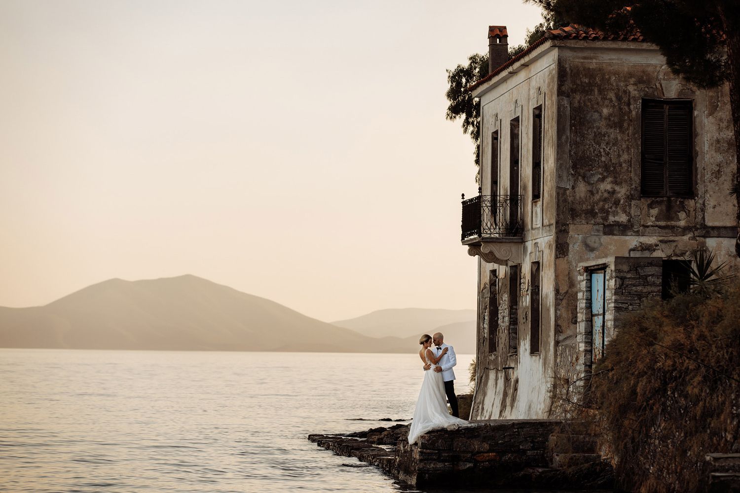 A romantic wedding photograph at sunset by an old stone villa overlooking a serene Mediterranean coastline and mountains.