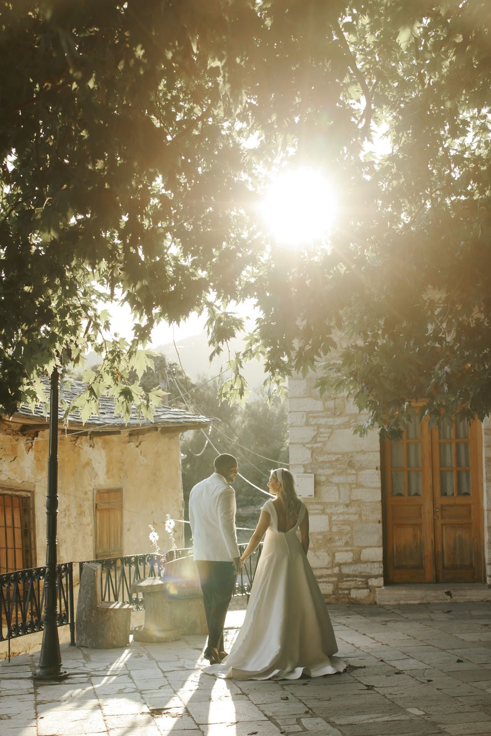 Bride and groom holding hands at golden hour, standing on stone patio surrounded by trees and rustic building.