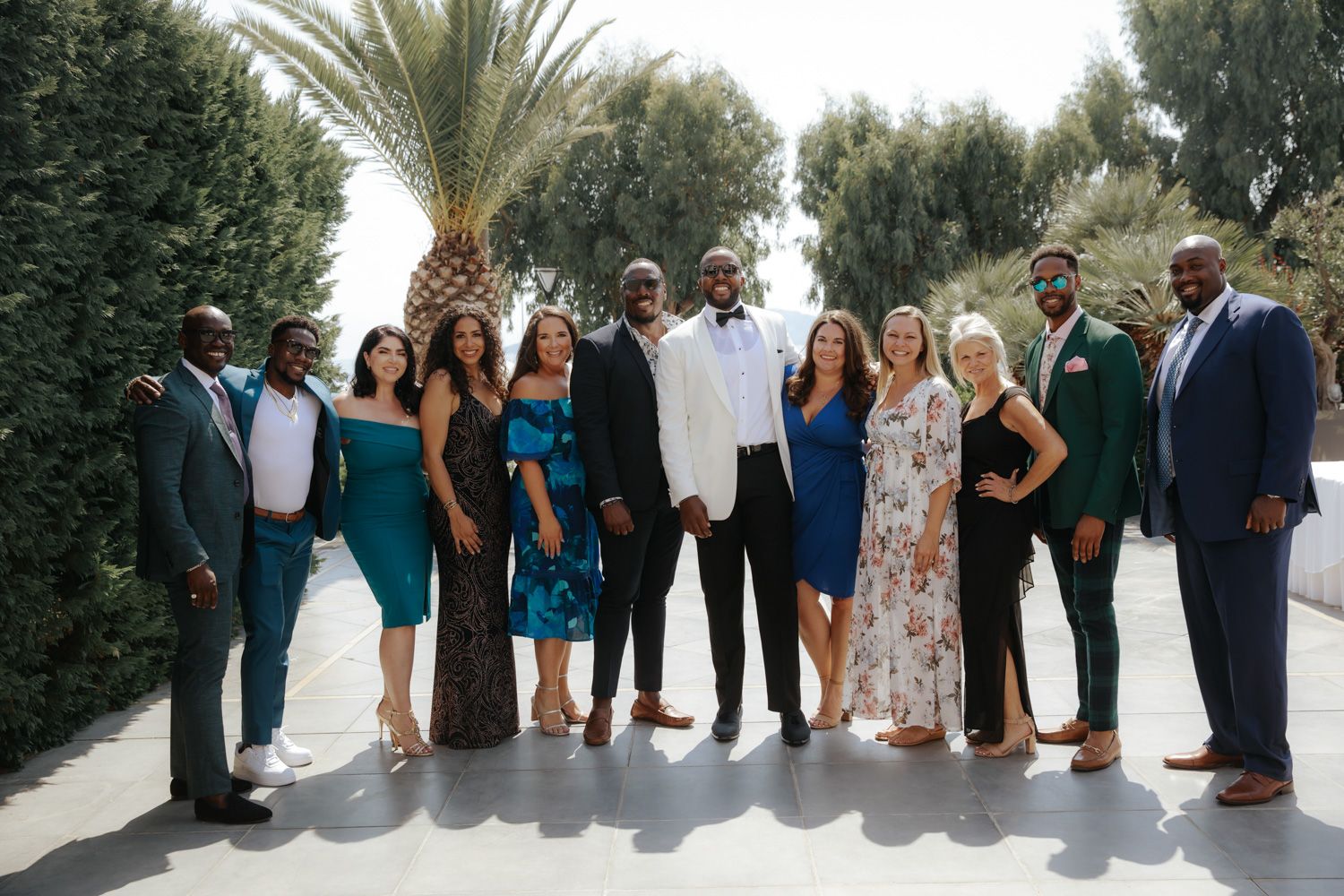 A diverse group of elegantly dressed friends posing together outdoors at a formal event near a palm tree.