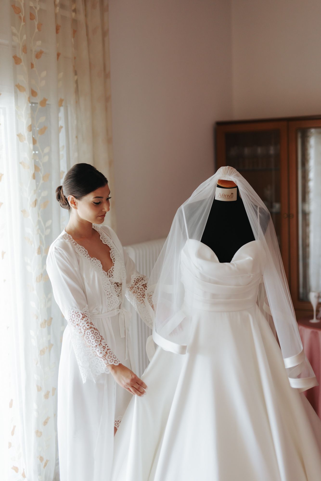 Bride in white lace robe admiring her elegant wedding gown on a mannequin near a bright window.