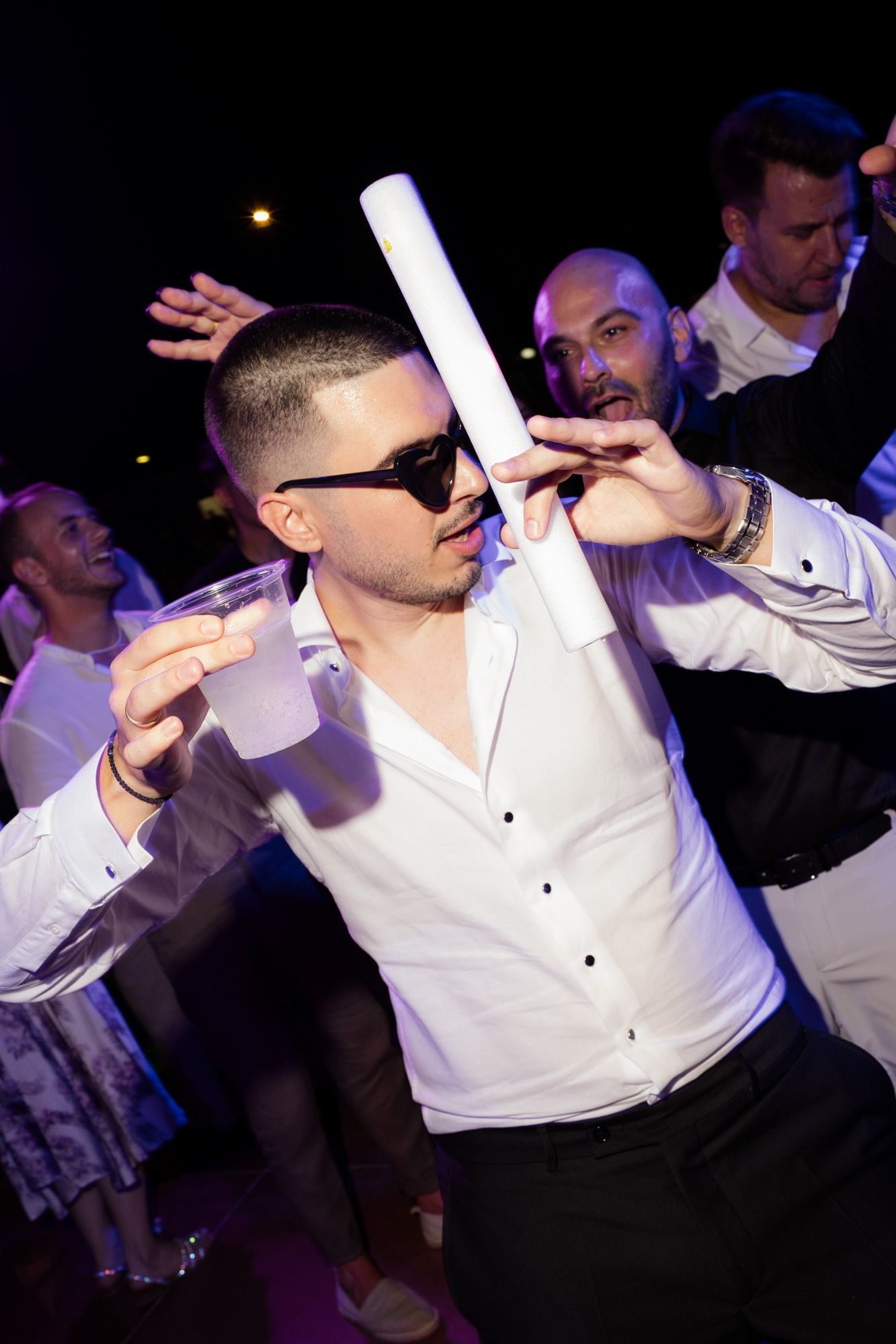 Man in white shirt and sunglasses dancing at a party, holding a drink and white tube under colorful lights.