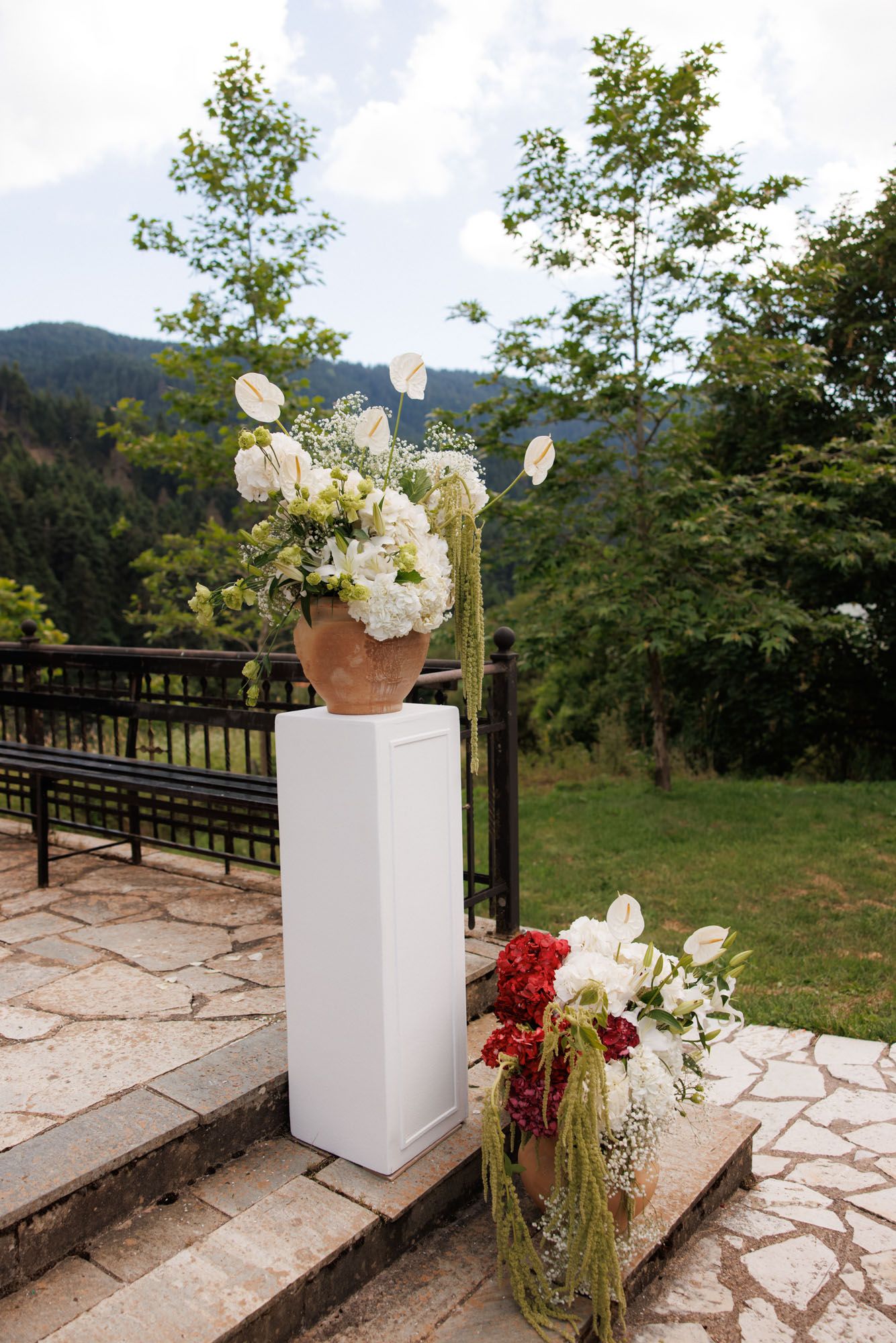 Elegant white floral arrangements in terracotta vase on pedestal with mountain forest backdrop at outdoor wedding.