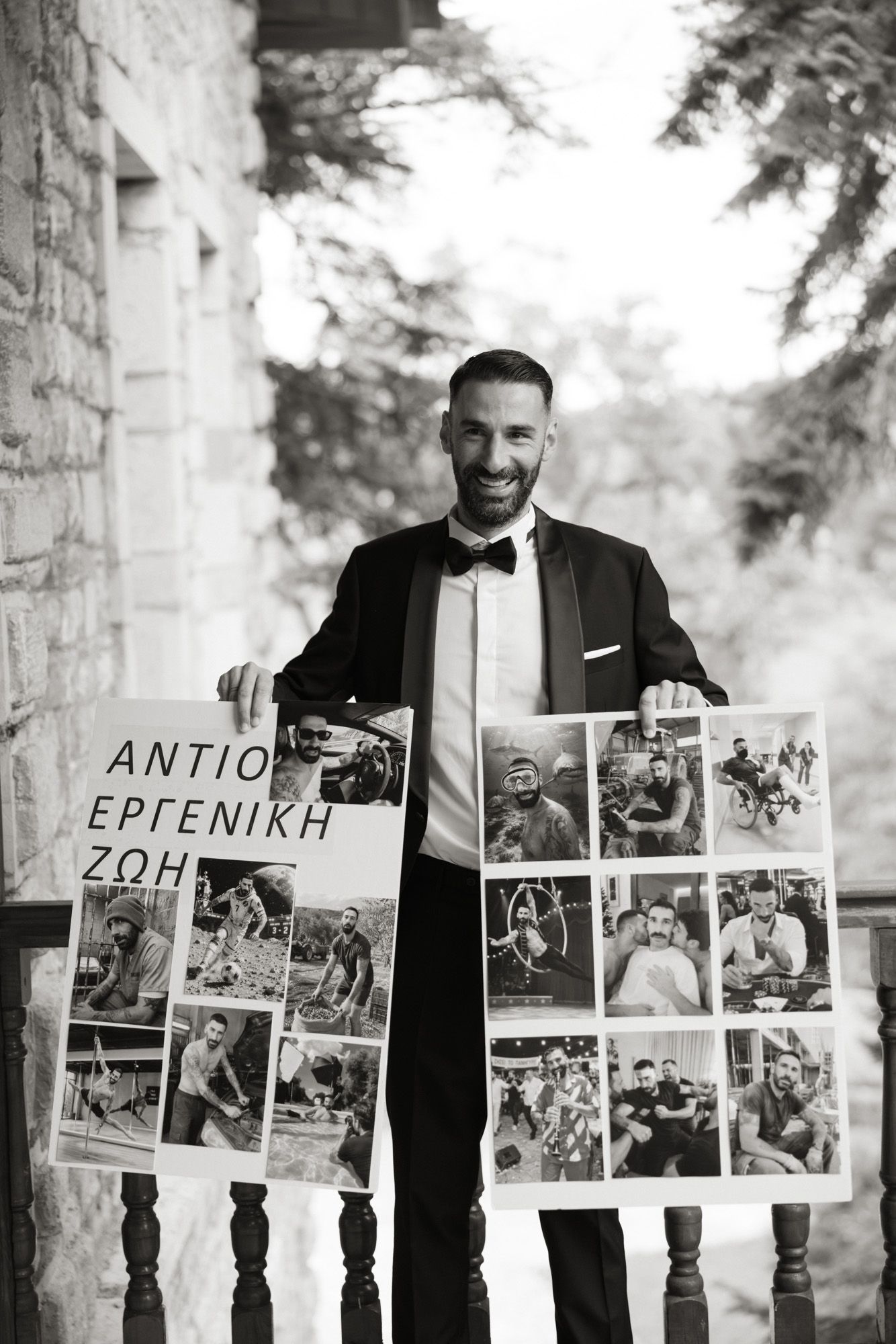 Smiling groom in tuxedo holds wedding photo collage boards with Greek text at outdoor venue.