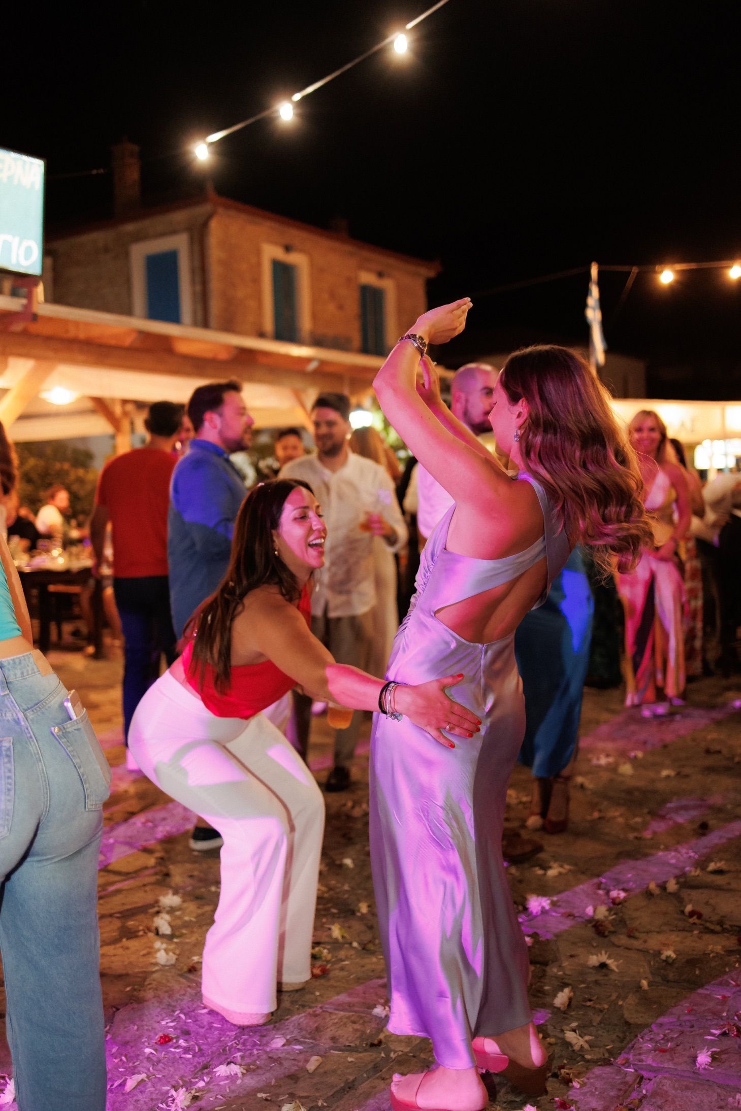 Two women dancing joyfully at an outdoor wedding reception under string lights at night.