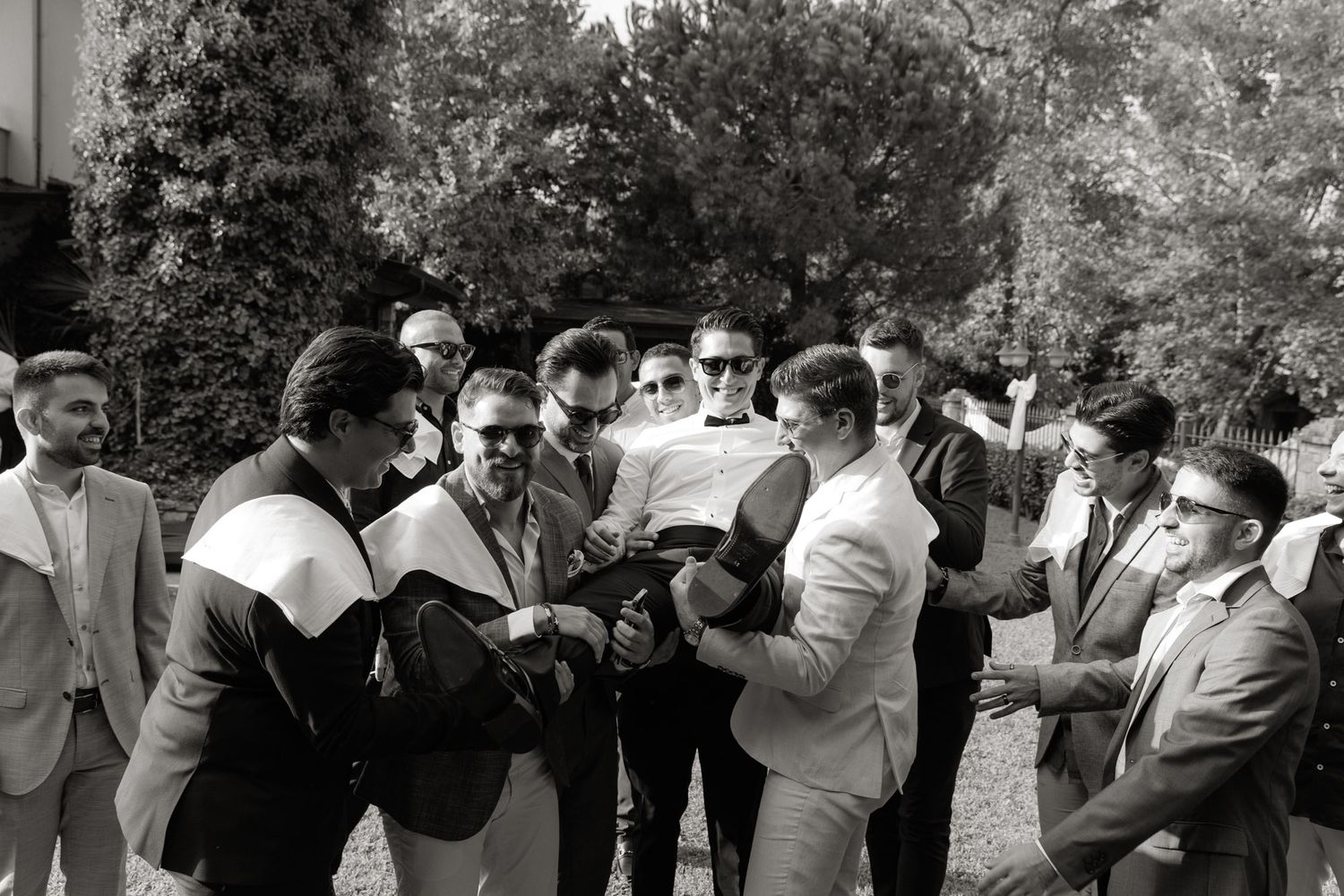 Black and white photo of groomsmen gathered together outdoors, laughing and celebrating before a wedding ceremony.
