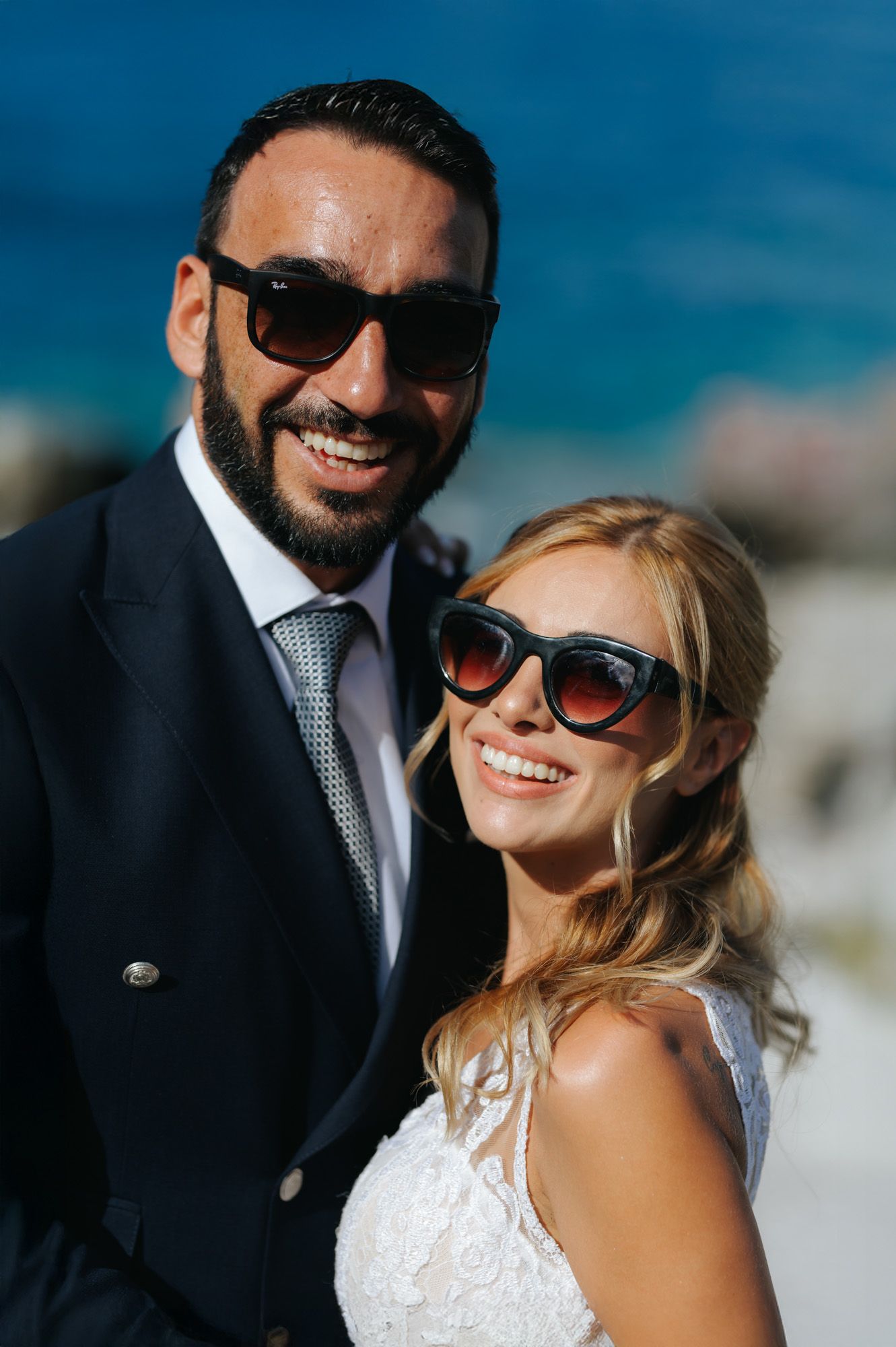 Smiling bride and groom wearing sunglasses pose together outdoors with blue sea in the background.