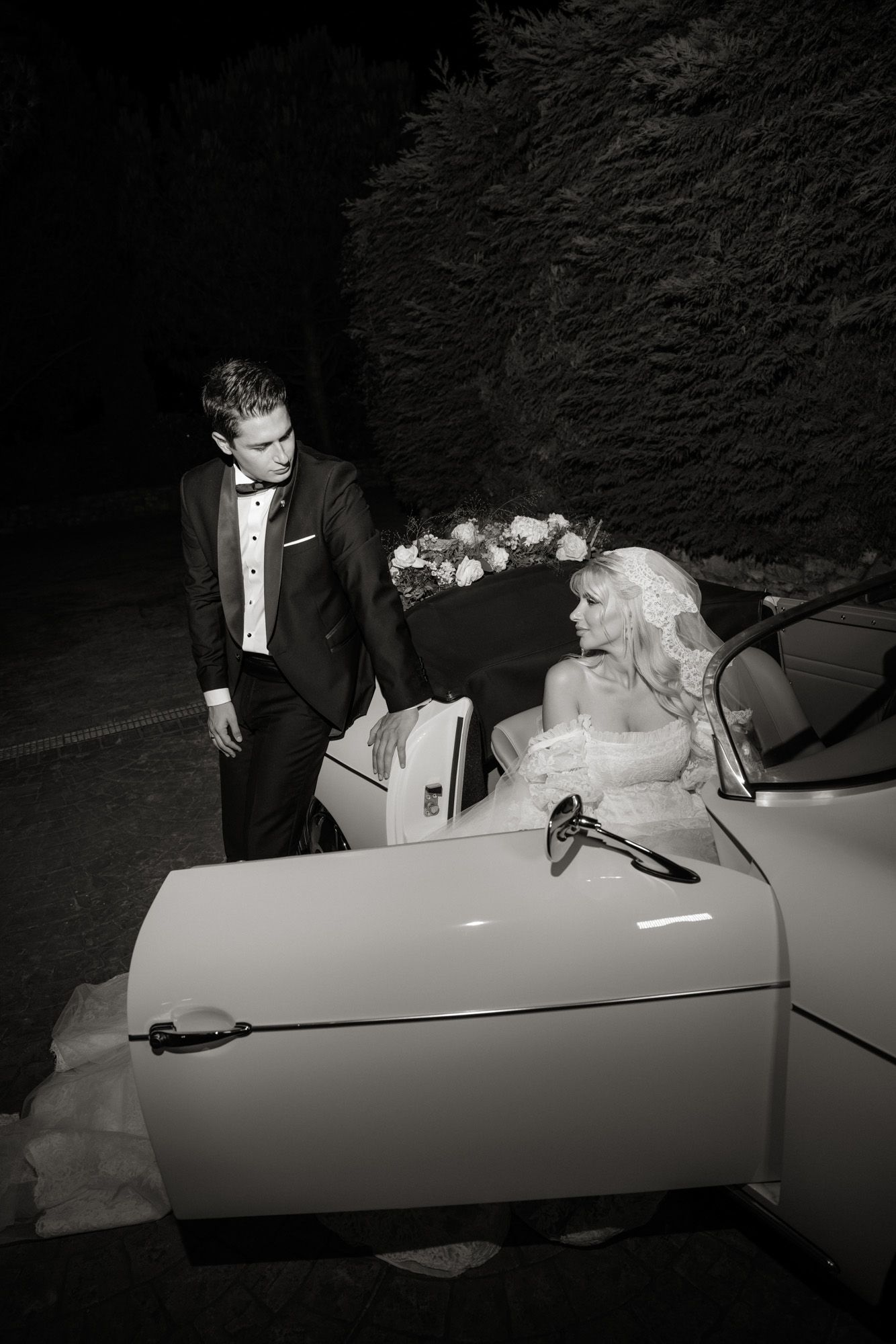 Black and white wedding photo of groom in tuxedo opening car door for bride seated in vintage convertible.