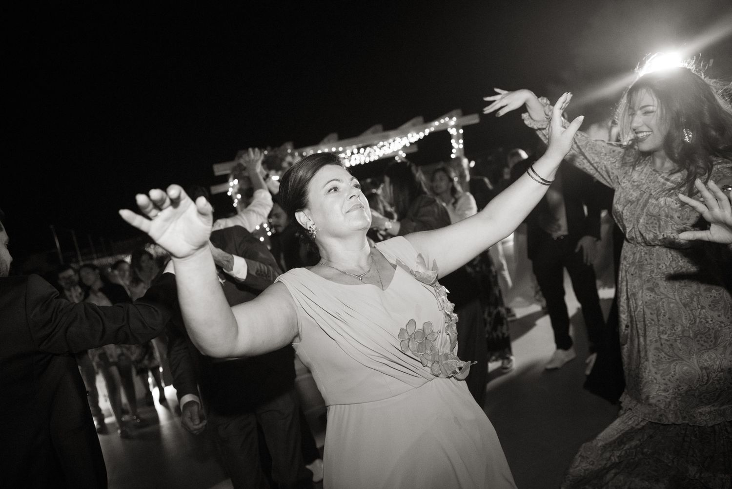 Black and white photo of a joyful woman dancing with arms outstretched at an outdoor wedding reception at night.