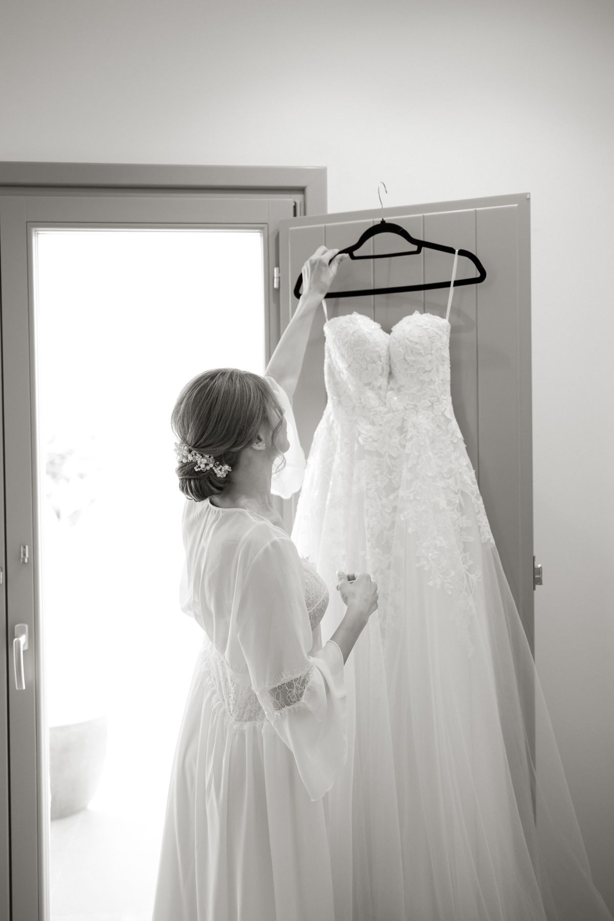 Bride in white robe admiring her lace wedding gown hanging on a door in black and white.