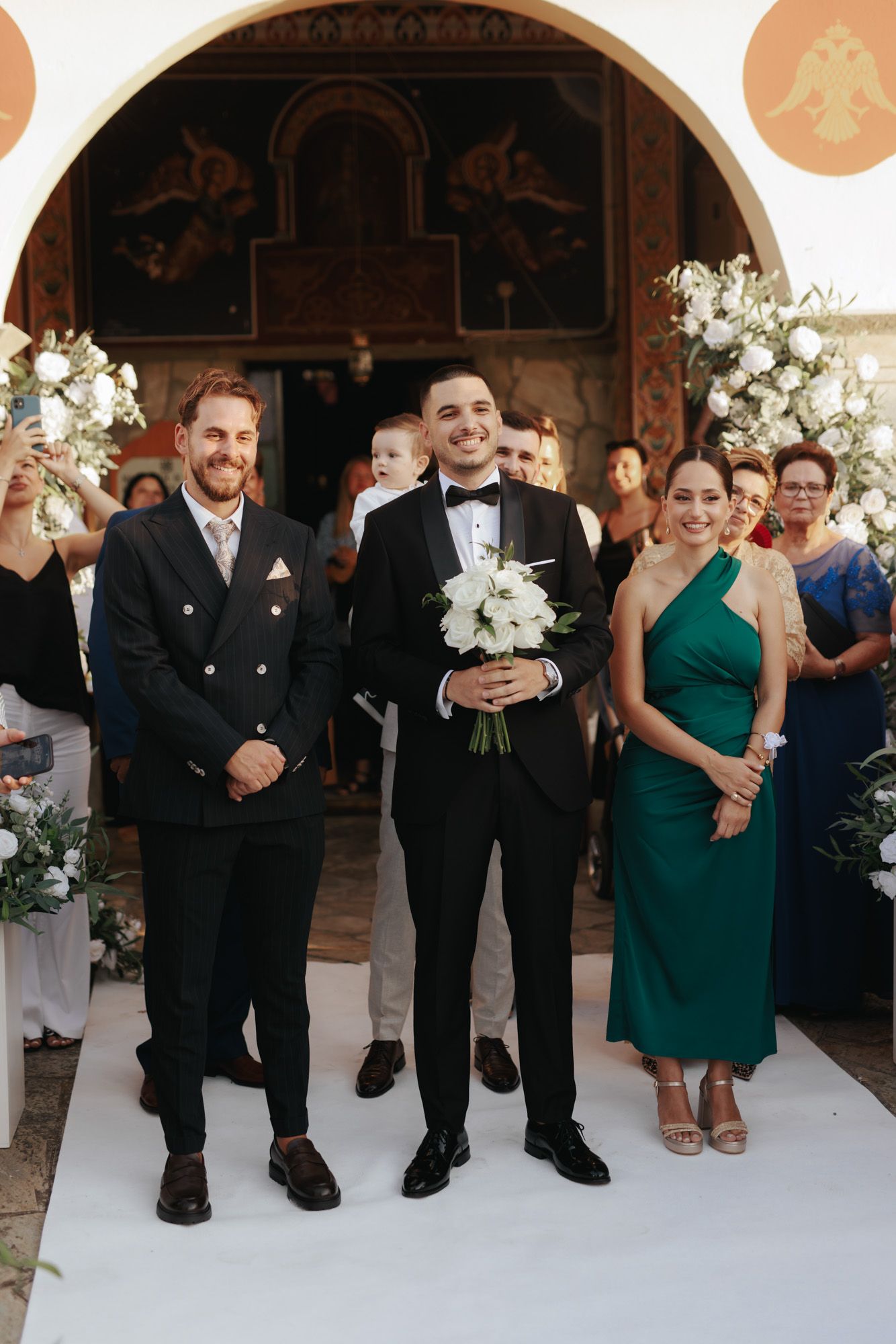 Groom in tuxedo walks down aisle holding white bouquet, flanked by smiling man in suit and woman in green dress.