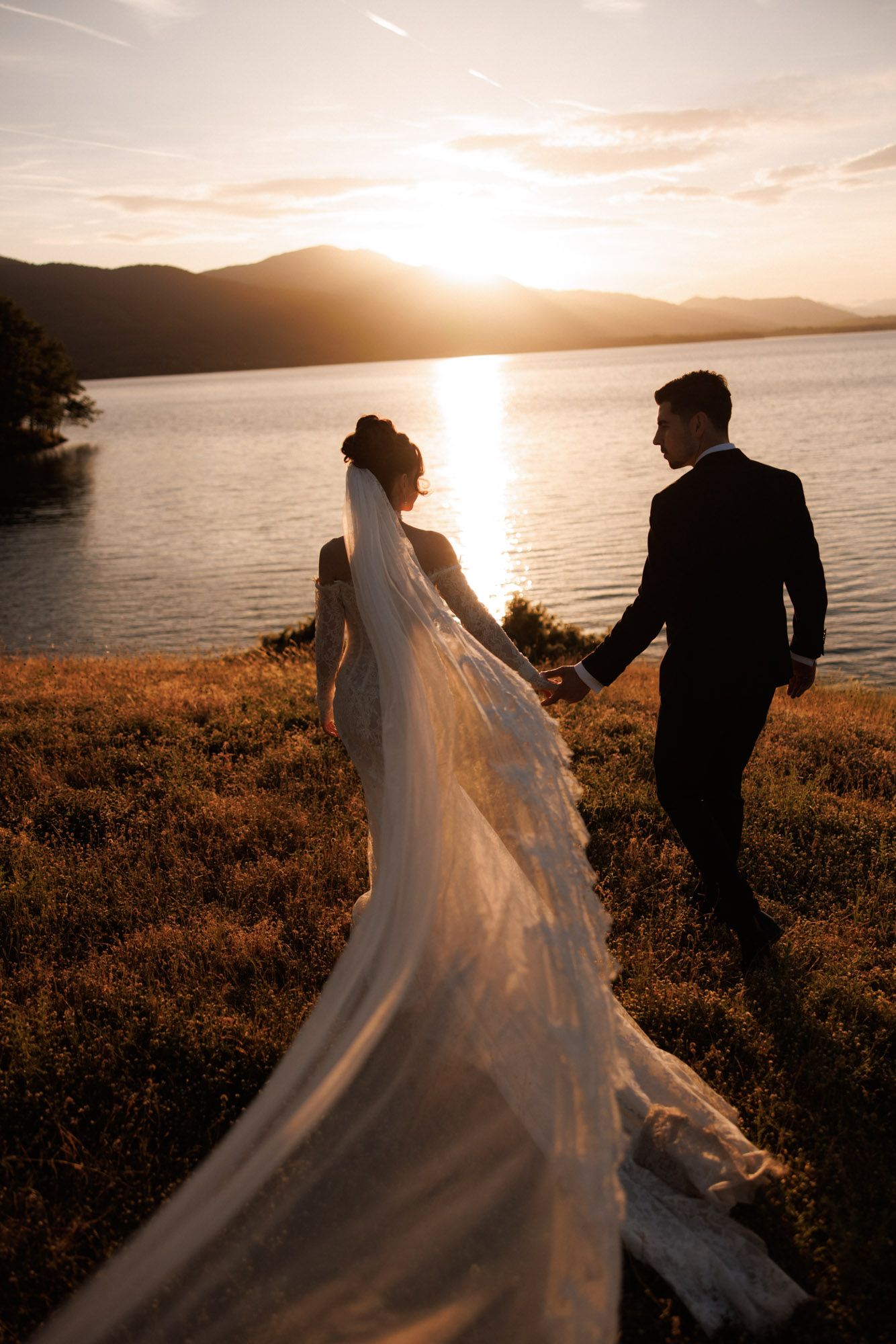 Bride and groom hold hands at sunset by a mountain lake, her long veil flowing behind in golden light.