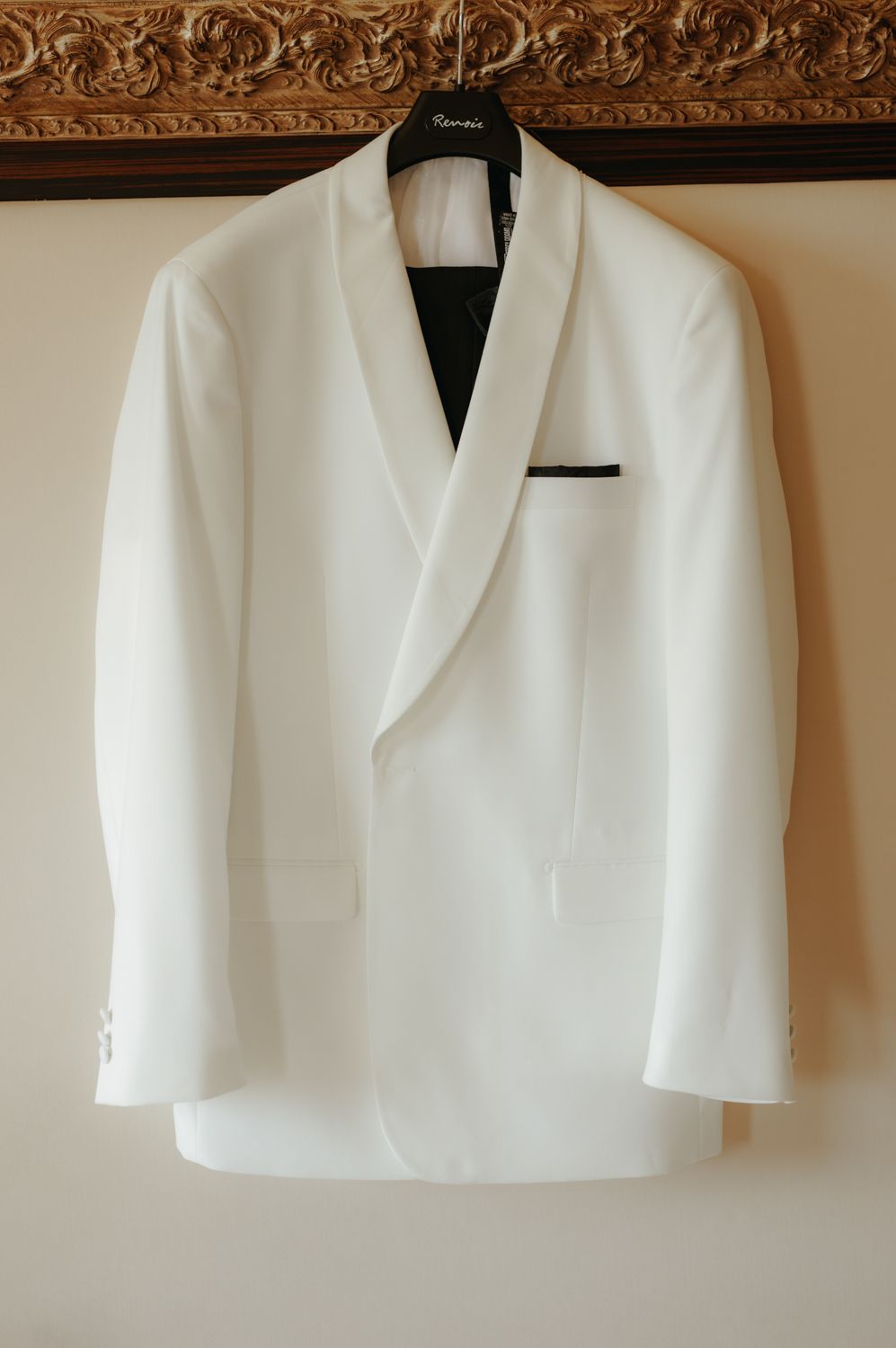 White double-breasted tuxedo jacket with black pocket square hanging on a hanger against a neutral wall.