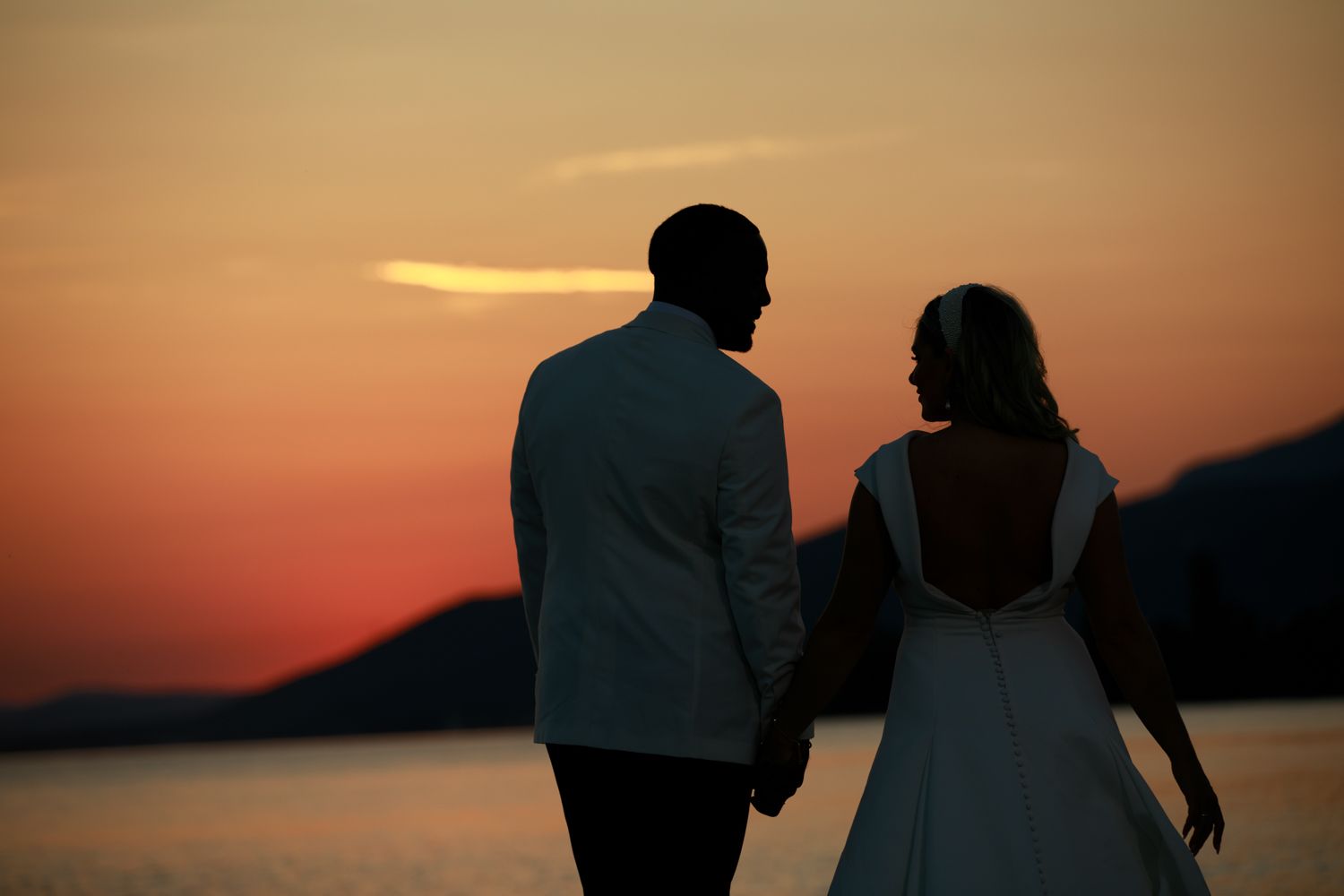 Couple in wedding attire holding hands watching a romantic sunset over the ocean with silhouetted mountains.