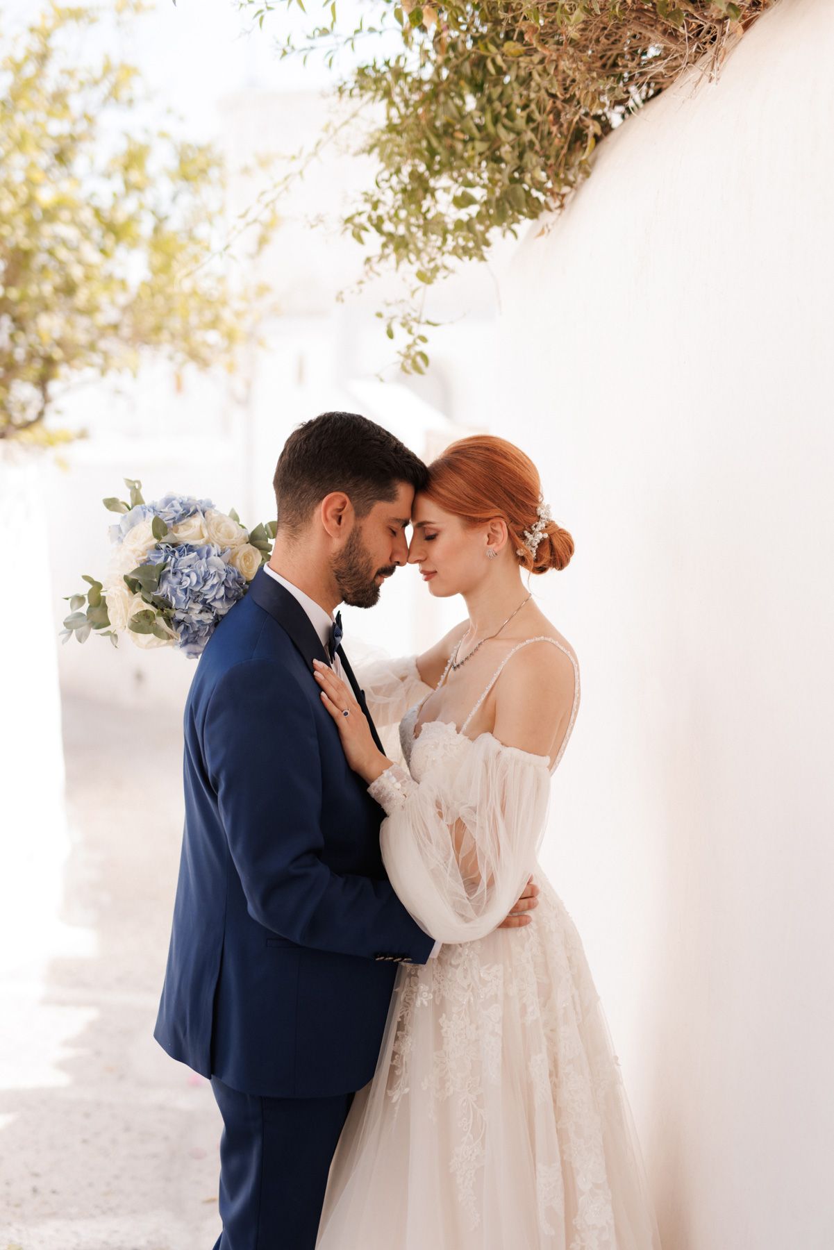 Bride and groom sharing an intimate forehead touch moment in a sunlit white alley with blue hydrangeas.