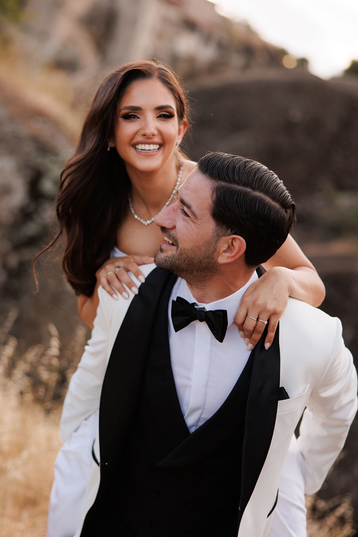 Happy bride and groom in elegant wedding attire sharing a joyful piggyback moment outdoors.